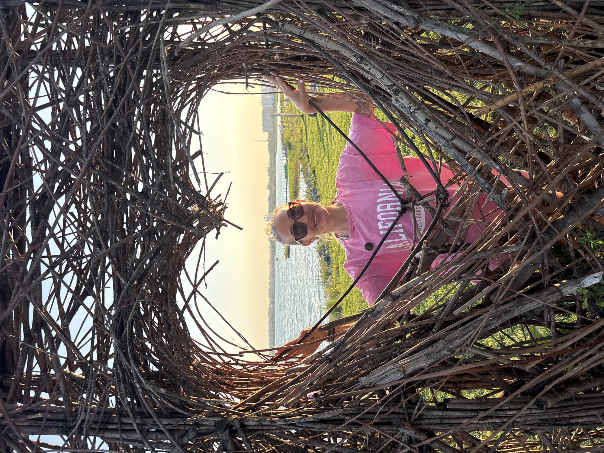 A woman stands within a large, heart-shaped sculpture made of intertwined branches, positioned near a body of water. The sculpture frames her, with branches extending in front of and behind her. She is wearing a pink sweatshirt with the word "CALIFORNIA" and "WEST COAST" printed on it, sunglasses, and has blonde hair pulled back. Her arms are extended, resting on the branches of the sculpture. The background shows a calm body of water, a grassy shoreline, and buildings in the distance under a hazy sky with a warm glow, suggesting either sunrise or sunset. The scene appears to be an outdoor recreational area.