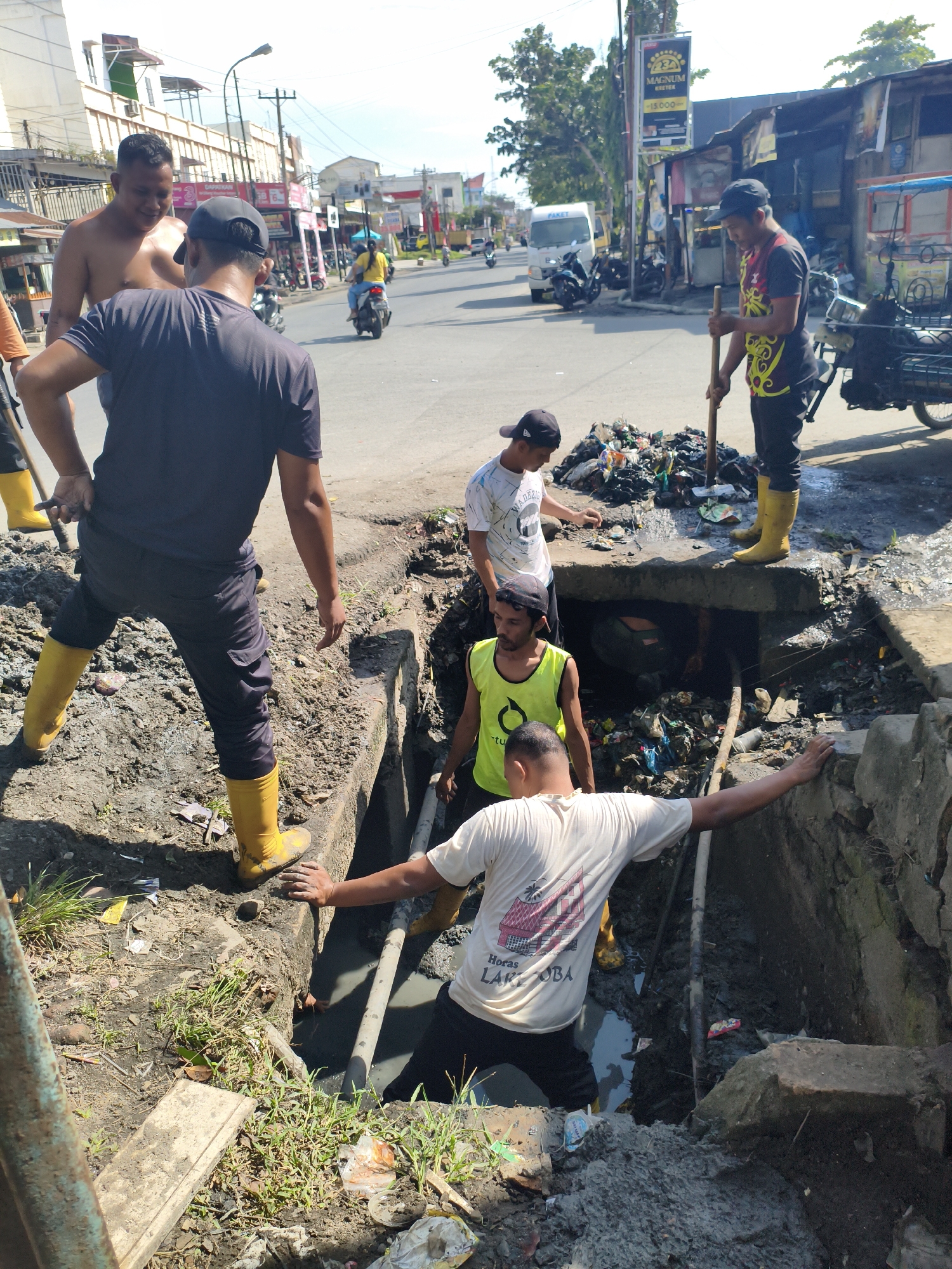 In the City of Tebing Tinggi, Indonesia, a group of men are engaged in clearing a clogged drainage system. The scene is a street with buildings and ongoing traffic. One man, shirtless and wearing black pants and yellow boots, stands in the foreground, leaning over the drainage trench. Another man, wearing a black t-shirt and black pants with yellow boots, is standing further down the street, holding a wooden stick and attending to a pile of garbage. A third man, in a white t-shirt and black pants, is also near the drainage. Two other men are partially visible, one wearing a yellow vest and the other in a white t-shirt. The drainage trench is filled with murky water and debris, indicating a significant blockage. The environment appears to be a typical urban street with a mix of commercial and residential structures. The overall impression is one of a community effort to address a sanitation issue.