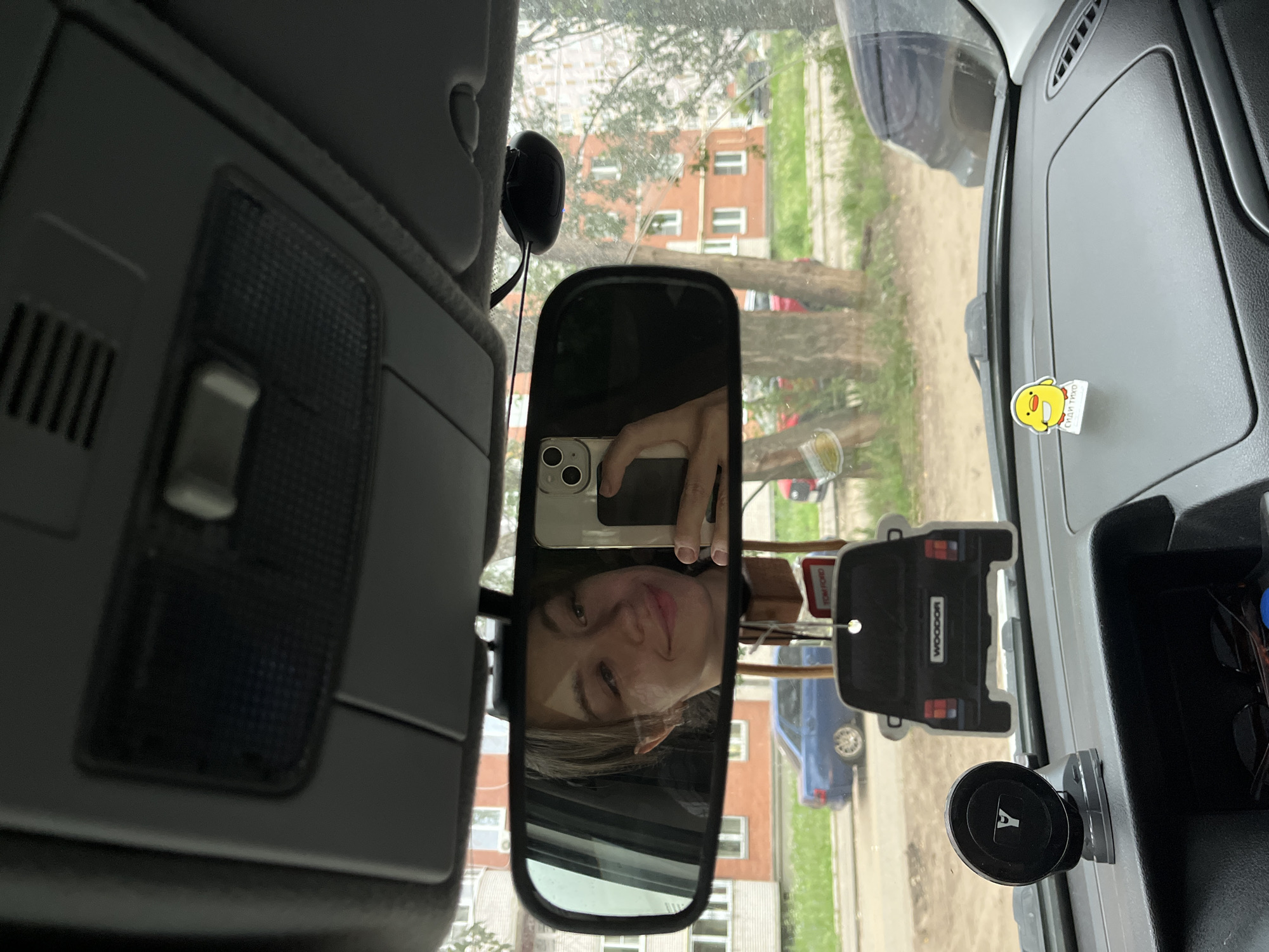 The image captures a reflection in a car's rearview mirror. A young woman is visible, holding a smartphone and taking a selfie. She has light brown hair styled loosely and is smiling at the camera. The reflection also shows the interior of the car and the exterior environment.
Outside the car, there are buildings, trees, and a blue car parked on the street. The architecture of the buildings suggests a typical urban or suburban setting in Russia, likely in Chaikovsky. The presence of a car and its surroundings implies the image was taken while the car was parked or in slow-moving traffic.
Inside the car, on the dashboard, there are several objects: a black suction cup mount, an air freshener shaped like the back of a car with "WOODOR" written on it, and a small decorative item resembling a yellow chick with text on its base, which appears to be Russian lettering. The overall scene is a casual snapshot taken by someone inside their vehicle.