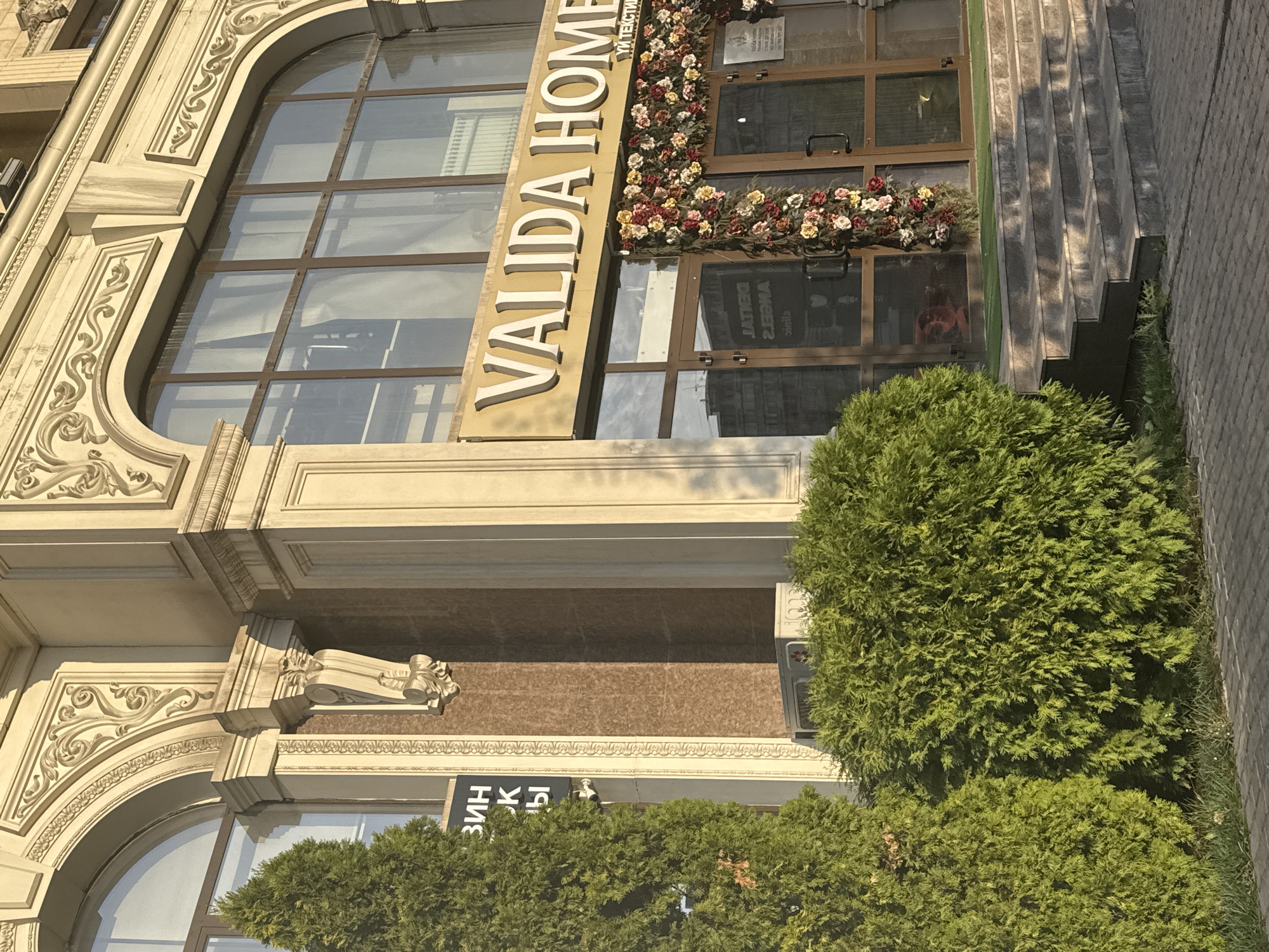 The image shows the entrance of a building with an ornate facade, likely a commercial establishment. The building features large, arched windows with a beige, classical-style decorative overlay. Below the windows, there are glass doors adorned with a garland of artificial flowers in red, yellow, and white. A sign above the doors reads "VALIDA HOME". To the left of the entrance, there are two coniferous trees and a bush. A sign with Cyrillic lettering is partially visible on the left side of the building. The entrance area has a set of stone stairs leading up to the doors, and the ground is paved with cobblestones. In the reflection of the glass doors, an urban environment with buildings can be seen. The lighting suggests it is daytime, with sunlight casting shadows. This establishment is located in Bishkek City, Kyrgyzstan.