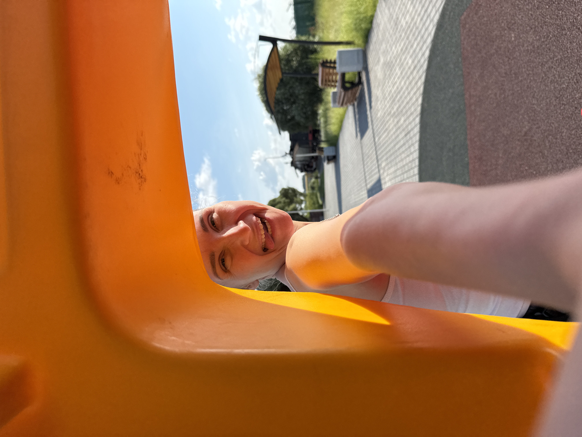 A woman is peeking through an orange plastic structure in a city park. She has blond hair and is wearing a white top. She is sticking her tongue out playfully. In the background, there is a paved walkway, benches, and some greenery. The sky is blue with some clouds. The woman's arm and hand are visible in the foreground, holding a camera or phone to take the picture.