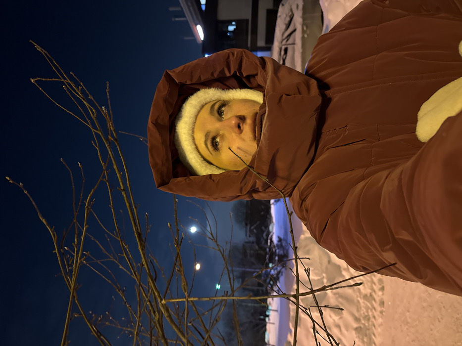 The image displays a close-up, self-portrait taken outdoors during the evening or night in a winter setting. The perspective is from slightly below, looking up towards the subject and the sky.
A person is centrally featured, wearing warm winter attire. They have a dark reddish-brown or maroon puffer jacket with the hood pulled up, encircling their head. Underneath the hood, a light-colored, possibly cream or white, knitted hat is visible, framing their face. Their eyes are looking directly at the camera, and their eyebrows and eyelashes are dark. The lower part of their face is mostly obscured by the jacket and hat. A thin, bare tree branch extends across the lower right side of their face. A light-colored, possibly fluffy or furry, material is visible on the upper right side of the frame, likely part of the jacket's design.
The background shows a deep blue, twilight or nighttime sky. To the left of the person, a network of bare tree branches, possibly with small buds, fills a significant portion of the foreground. In the mid-ground, a prominent, circular bright light, resembling a full moon or a strong distant light source, glows in the sky. Below this light, other smaller, blurred artificial lights are visible on the horizon, hinting at a distant urban or suburban area. The ground is covered in a blanket of snow, reflecting some of the ambient light, and appears as a bright, uneven surface stretching into the background. Hints of structures or buildings with lights can be seen in the far background on the right.
The overall impression is one of cold weather, with the person bundled up warmly against the elements. No text is visible in the image.
