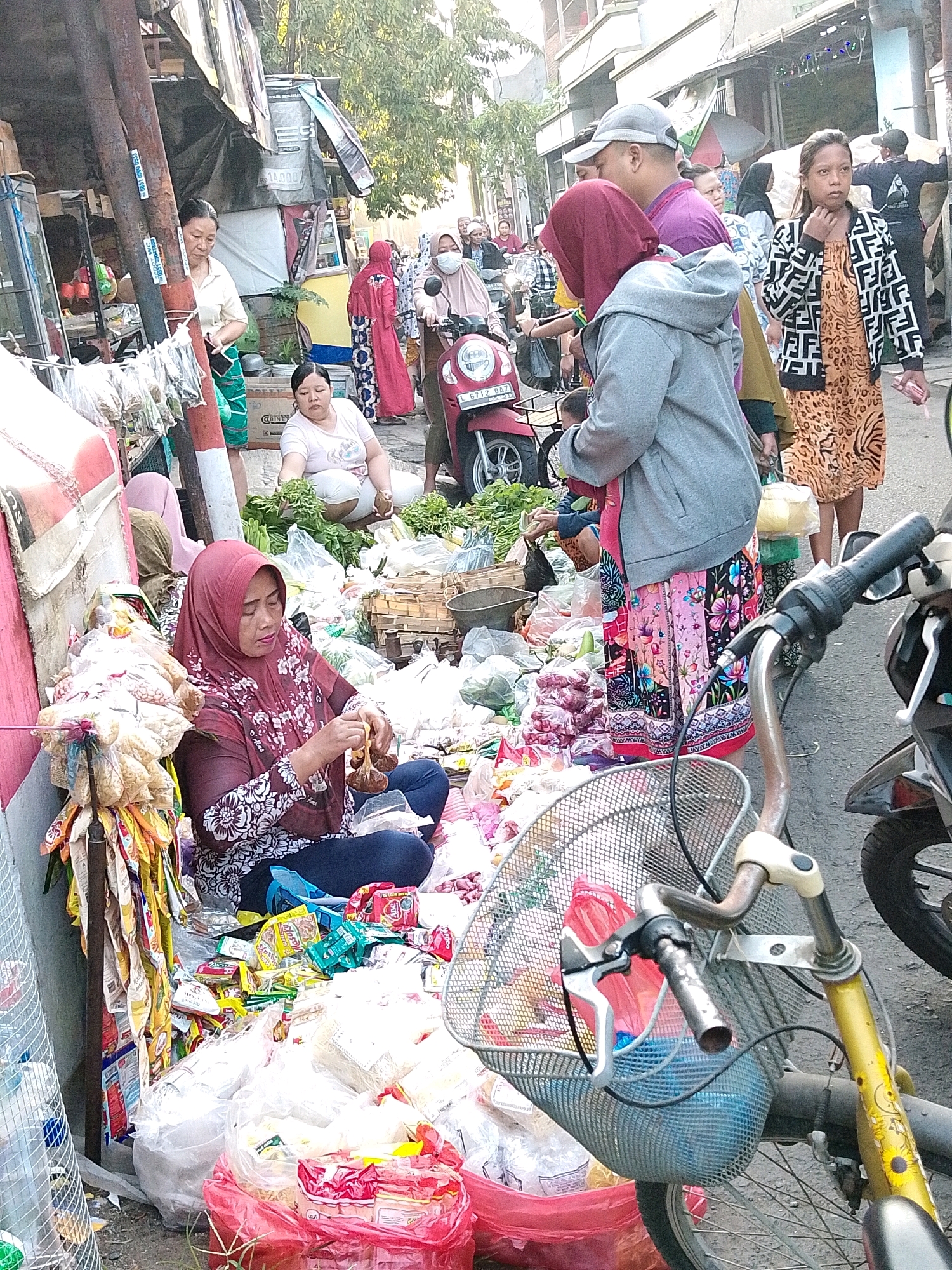 The image depicts a bustling street market scene in Surabaya, Indonesia. In the foreground, a woman wearing a maroon hijab and a floral print blouse sits on the ground, sorting through small, brown, bulb-like items in her hands. She is surrounded by a large assortment of goods, including plastic bags filled with various food items, some appearing to be packaged snacks and others possibly produce like onions or garlic. A bicycle with a wire basket is positioned in the lower right corner, its handlebars and front wheel visible.
In the background, other individuals are present. To the left, a woman in a cream-colored shirt and green skirt stands near a stall with hanging items. Further back, a woman wearing a pink hijab and a face mask is on a scooter, facing left. Other market stalls and shoppers are visible, with awnings, signage, and a variety of merchandise contributing to the busy atmosphere. The ground is paved and appears to be a street or alleyway, lined with buildings and trees in the distance, indicating an urban environment. The overall impression is one of a lively and crowded marketplace.