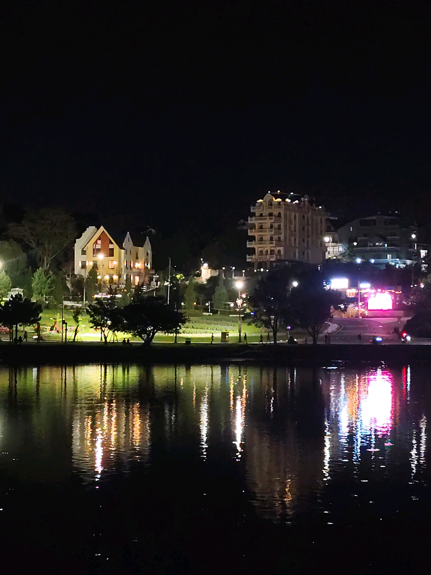 This is a nighttime photograph of Xuan Huong Lake in Da Lat, Vietnam. The lake's surface reflects the lights of buildings and streetlights on the shore, creating a vibrant display of colors against the dark water. On the left side of the image, two buildings with unique architecture, possibly a hotel or resort, are illuminated. To the right, a taller, modern building with multiple floors is visible, along with bright, colorful lights that suggest an entertainment venue or shops. The shore is lined with trees and a pathway where a few individuals can be seen walking. The overall atmosphere is serene and picturesque, typical of Da Lat's popular tourist spots after dark.