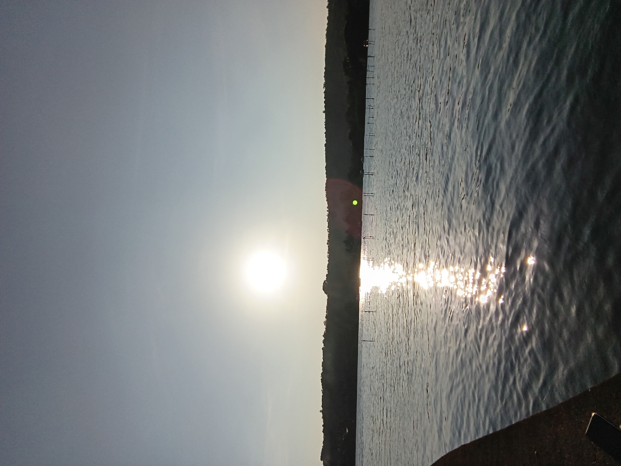 The image depicts a body of water reflecting sunlight, with a treeline visible on the opposite shore. The sun is positioned prominently in the sky. Wooden poles are present in the water near the shoreline. Part of a structure is in the foreground on the bottom right. No people are visible in the image. The location is specified as Oxel, India, but there's no visible landmarks or evidence to confirm this.