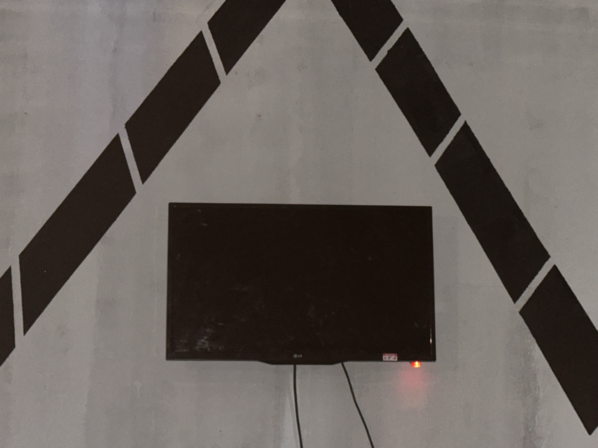 The image captures a wall with a decorative black geometric pattern resembling an inverted "V" or an arrow pointing downwards, with dashed lines. In the center of the wall, a flat-screen television is mounted. The television is off, but a small red indicator light is visible on its lower right side, suggesting it is in standby mode. The wall itself appears to be made of concrete and has a textured, somewhat rough surface, with some smudges and marks, indicating it might be an interior or exterior wall. The lighting suggests it is daytime, though the exact time of day is unclear due to the indoor or shaded setting. The overall scene is static and depicts a typical domestic or public space where a television is displayed on a wall. There is no visible text in the image except for a small logo on the television, which appears to be "LG".