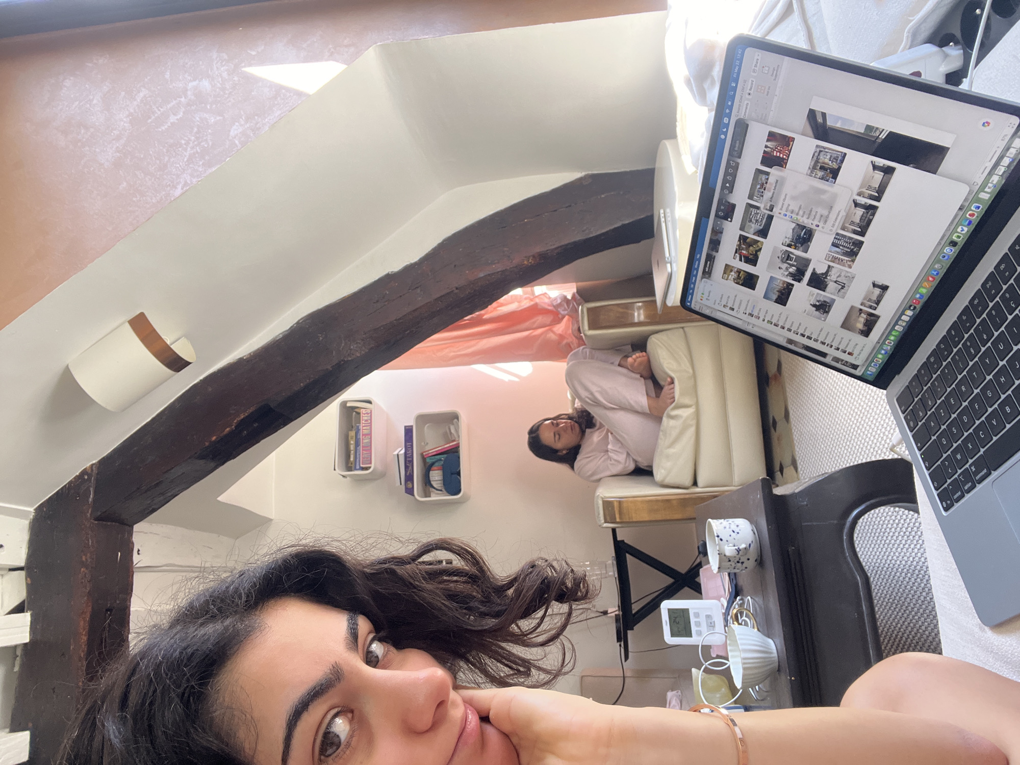 Here is a detailed description of the image:
**Setting:** The image appears to be taken inside a room, likely an apartment, characterized by a unique architectural design with exposed wooden beams and angled walls, indicating a possible attic or loft space. Bookcases are mounted on the wall.
**People:**
* A woman is the primary subject, partially visible in the foreground. She has dark hair and is looking directly at the camera. Her hand is visible near her face, possibly holding a phone or camera for a selfie.
* Another person is lying on a chaise-lounge, wearing pajamas, facing the laptop.
**Objects:**
* A laptop is prominently displayed, showing a collage of images.
* Two mugs, a device that looks like a remote control, and various small items are on a small table near the chaise-lounge.
* Books are arranged in shelves.
**Actions and Interactions:**
* The woman is taking a selfie, engaging with the camera.
* The second person appears to be relaxed on a chaise-lounge, possibly resting.
**Environment:**
* The room has white walls and light-colored decor, creating a bright and airy feel. The presence of the wooden beams adds a rustic element to the space.
* The setting likely suggests a residential apartment.
**Location in Paris:** While not explicitly visible, the style of architecture, notably the exposed beams and angled walls, is common in Parisian buildings, especially in older neighborhoods. Therefore, it's plausible the image was taken in Paris, France.