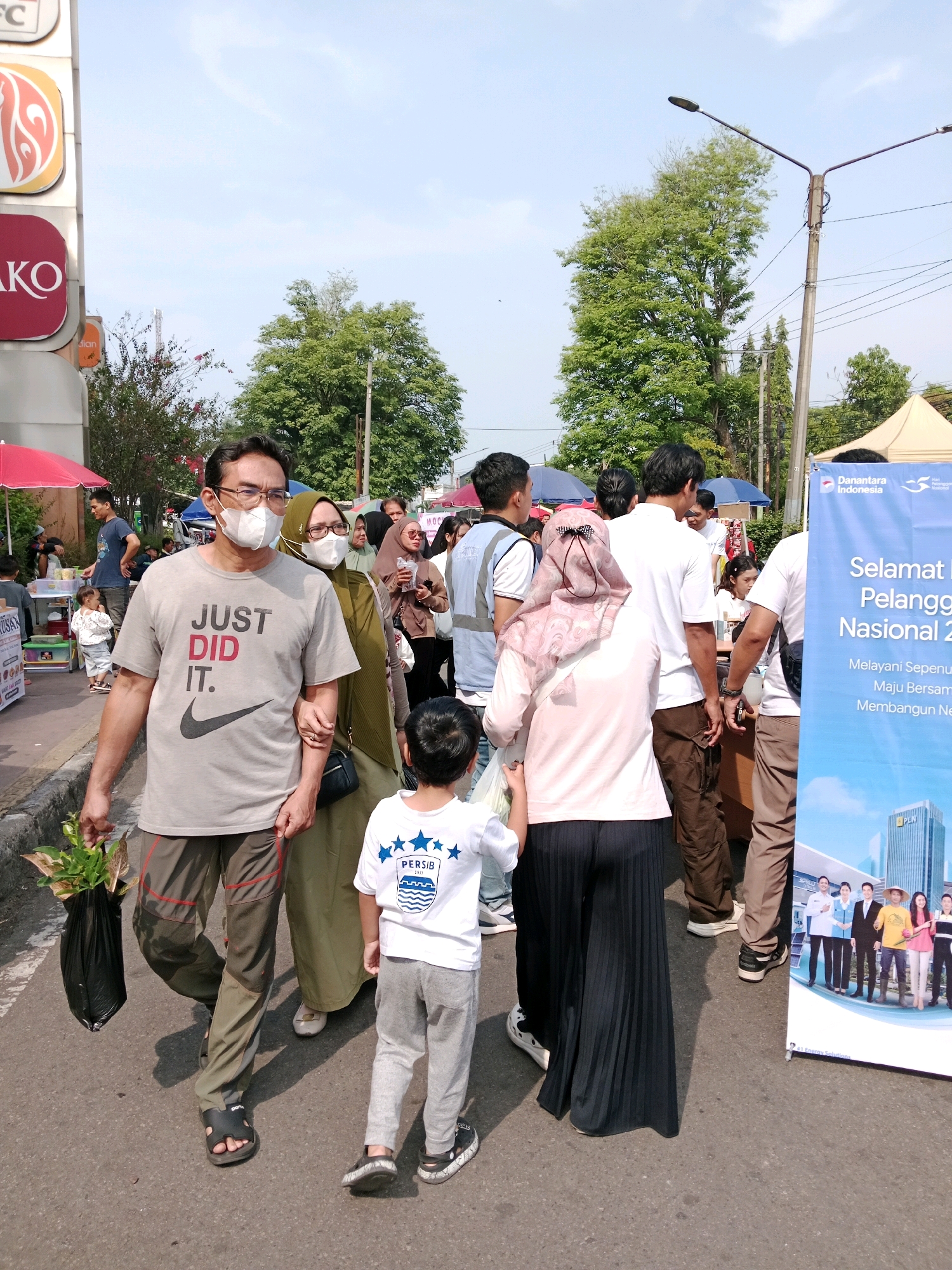 This image captures a street scene in Cianjur, Indonesia, likely during an outdoor event or market. In the foreground, a man wearing a gray t-shirt with the text "JUST DID IT." and glasses is walking with his arm linked with a woman in a green headscarf and glasses. Both are wearing face masks. A young boy in a white t-shirt with "PERSB" written on it walks ahead of them. The man is carrying a black plastic bag filled with plants. In the background, other people are visible, some under tents and umbrellas, suggesting a bustling environment. Signs and banners, including one with text in Indonesian, are present. Tall trees line the street, and a lamppost is visible. The ground is paved asphalt.