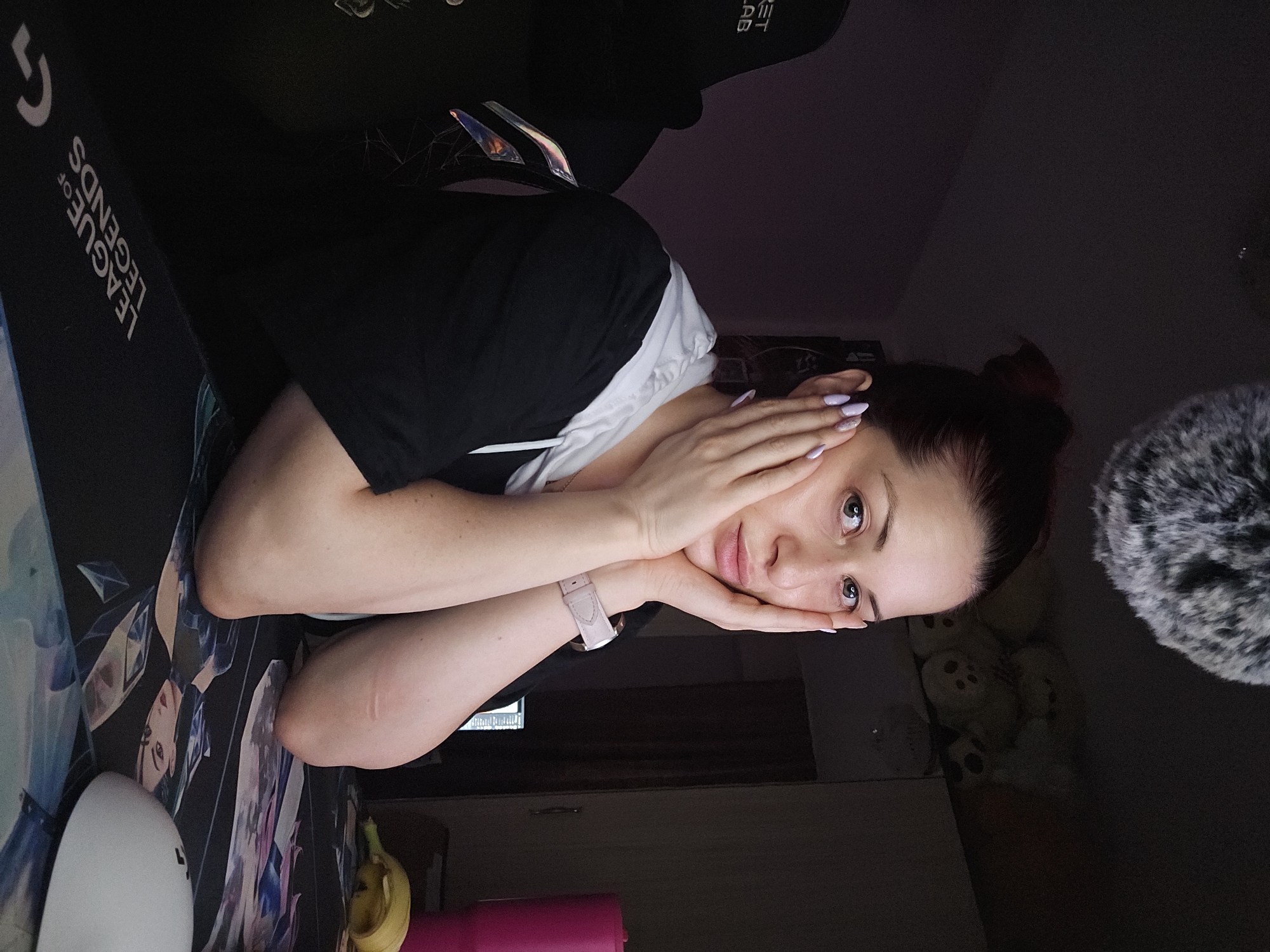 A young adult woman is depicted in a casual, indoor setting, resting her chin and cheeks on her hands while looking directly at the camera. Her dark hair is tied back, and she has light-colored eyes. She is wearing a black t-shirt over a white top, and her fingernails are painted a light lavender. A light pink smartwatch is visible on her left wrist.
The scene suggests a gaming or home office environment, dimly lit, likely in the evening or at night. The woman is positioned next to a large black desk mat featuring the text "LEAGUE OF LEGENDS" and stylized character art. On the mat, to her left, are a white oval-shaped device (possibly a lamp), a partially eaten banana, and a pink bottle or cup. Behind her, a black gaming chair with "LOGITECH" and "G" logos, along with "SECRET LAB" text, is visible. A headset with iridescent ear cups rests on the chair. To the right of the woman, a fluffy grey microphone wind muff is partially seen. In the background, several light-colored stuffed toys are dimly visible on a surface. The woman's expression is neutral and calm, portraying a relaxed moment. The location context provided is Burgas, Bulgaria.