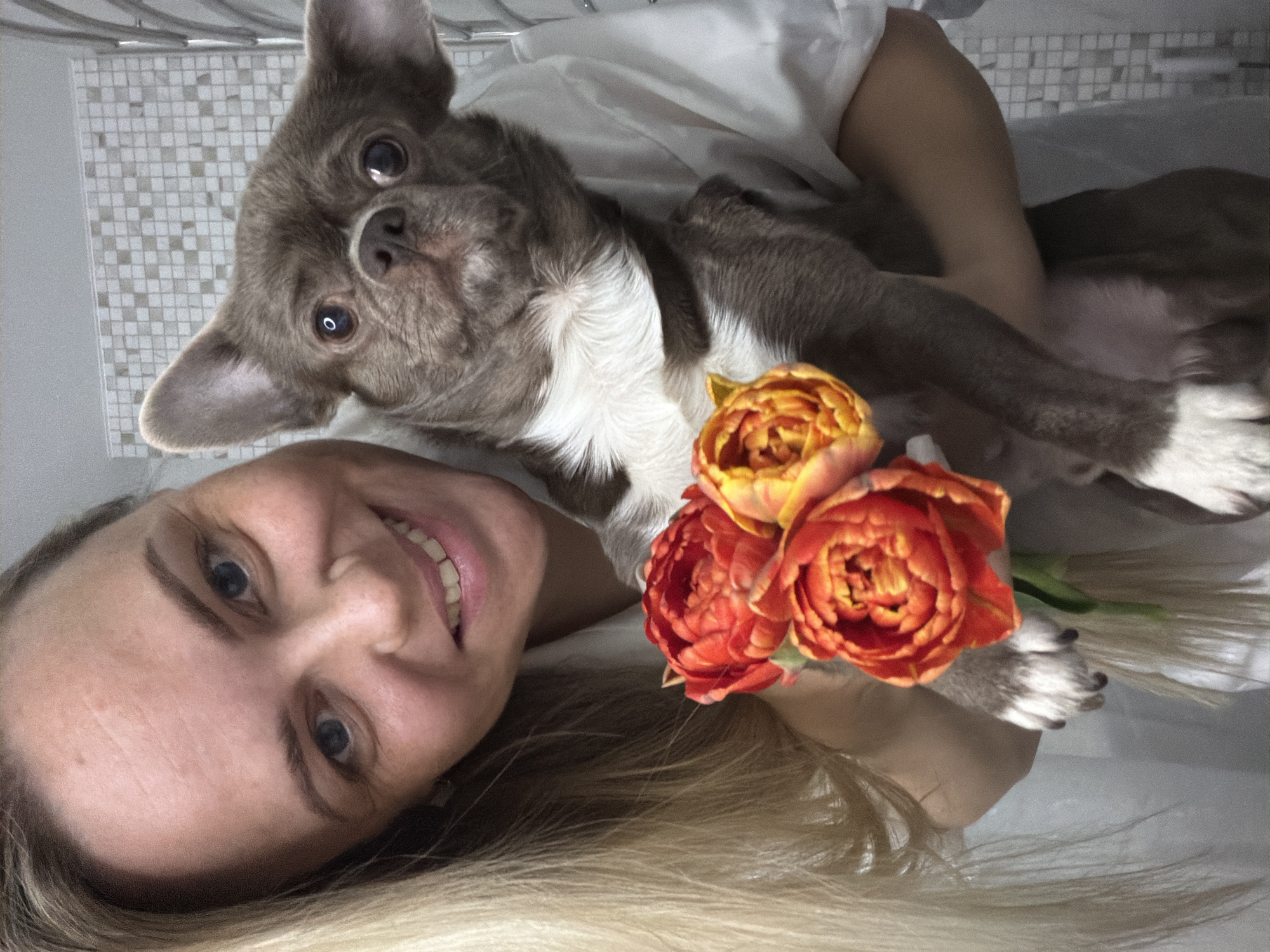 A woman is lying down, smiling at the camera, with a gray and white dog on top of her. She is holding three orange and yellow flowers in her hand. The background appears to be a tiled wall, possibly in a bathroom. There is no information to suggest that this image was taken in Votkinsk, Russia.