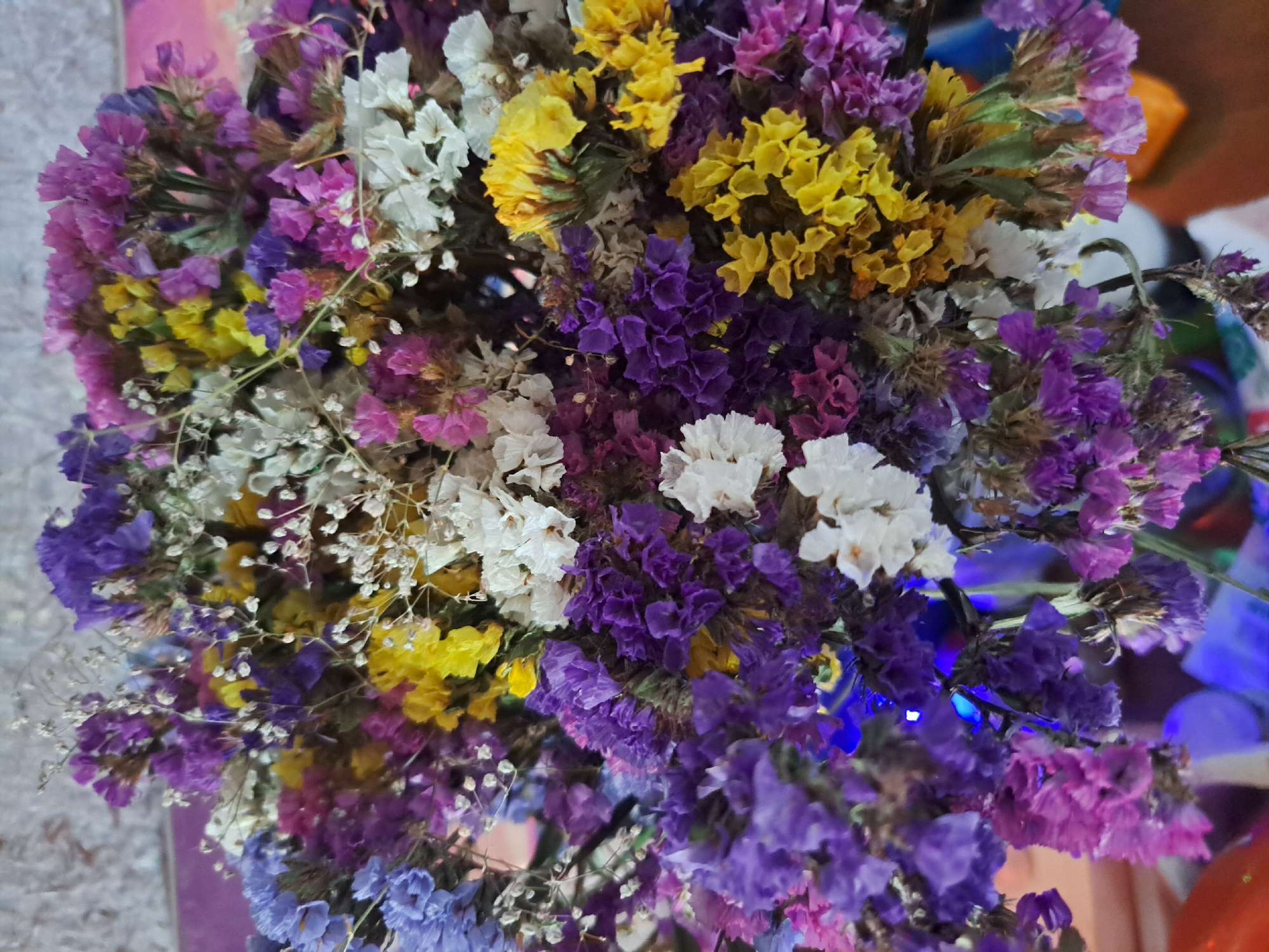 The image shows a close-up, overhead view of a bouquet of dried flowers. The flowers are in various shades of purple, yellow, blue, and white, with small, delicate petals. Some of the flowers are in full bloom, while others are more wilted and dried. The bouquet is arranged in a vase, the bottom of which is visible, with blue lights reflecting off its surface. The background is out of focus and appears to be a textured wall. There are no people or actions depicted in the image, and the scene is static. The location of this bouquet in Vypasne, Ukraine, cannot be determined from the visual information provided.