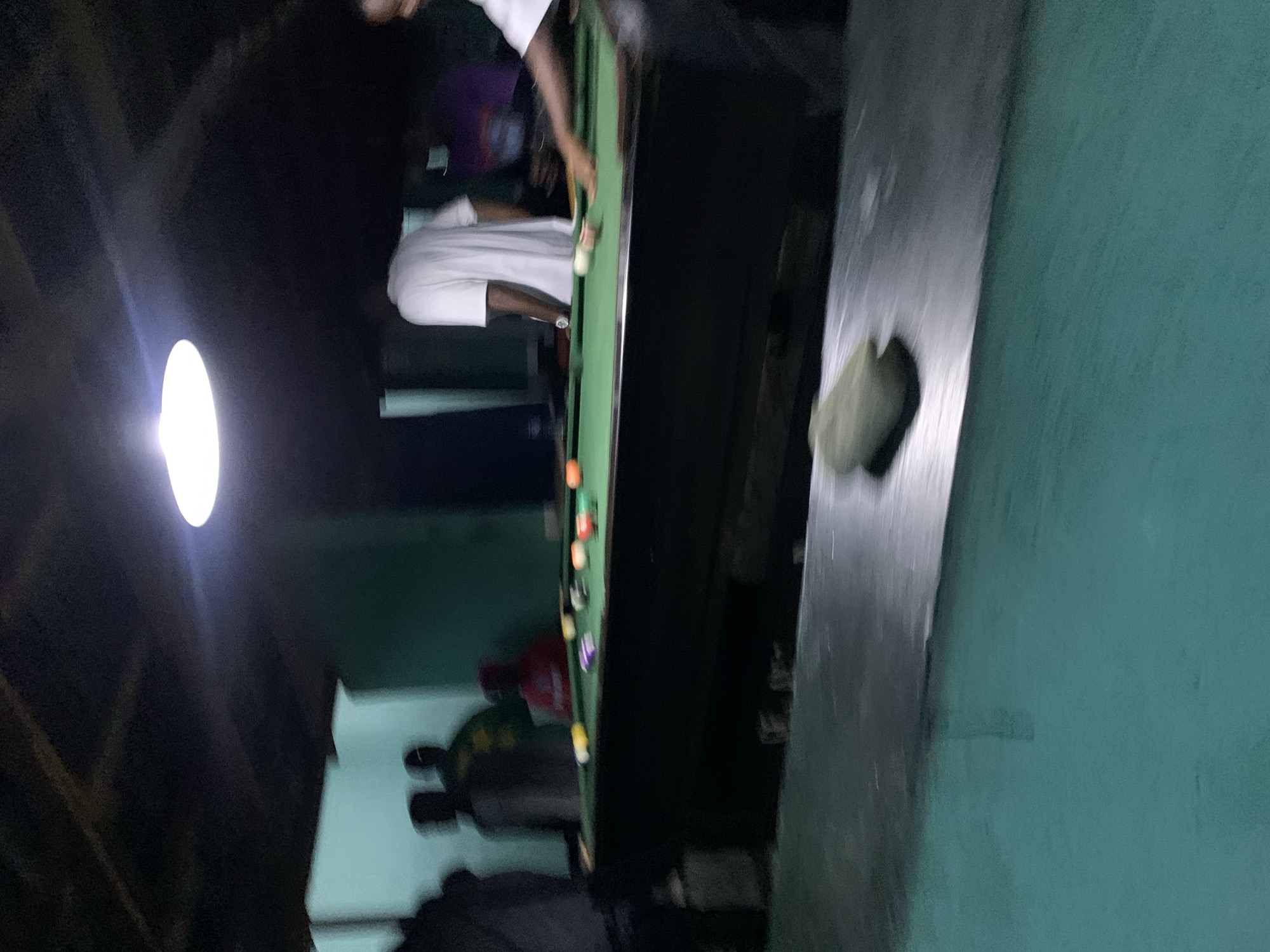 The image depicts an indoor recreational space in New Stadium, Nigeria, illuminated primarily by a bright overhead circular light source. A green-felt pool table occupies the midground, featuring several colored pool balls, including solids, stripes, and a cue ball. On the right side of the table, an individual in a white shirt is positioned, leaning forward with a pool cue, actively engaged in playing. Another person, also in a white shirt, stands on the opposite side of the table, facing away from the camera. In the background, multiple blurred figures are visible; one wears a red shirt, and another a yellow/green shirt. The room features light teal/green walls and a similarly colored floor. The ceiling appears dark and textured. In the immediate foreground, a dark, reflective surface is present, with a light-colored, irregular object resting upon it.