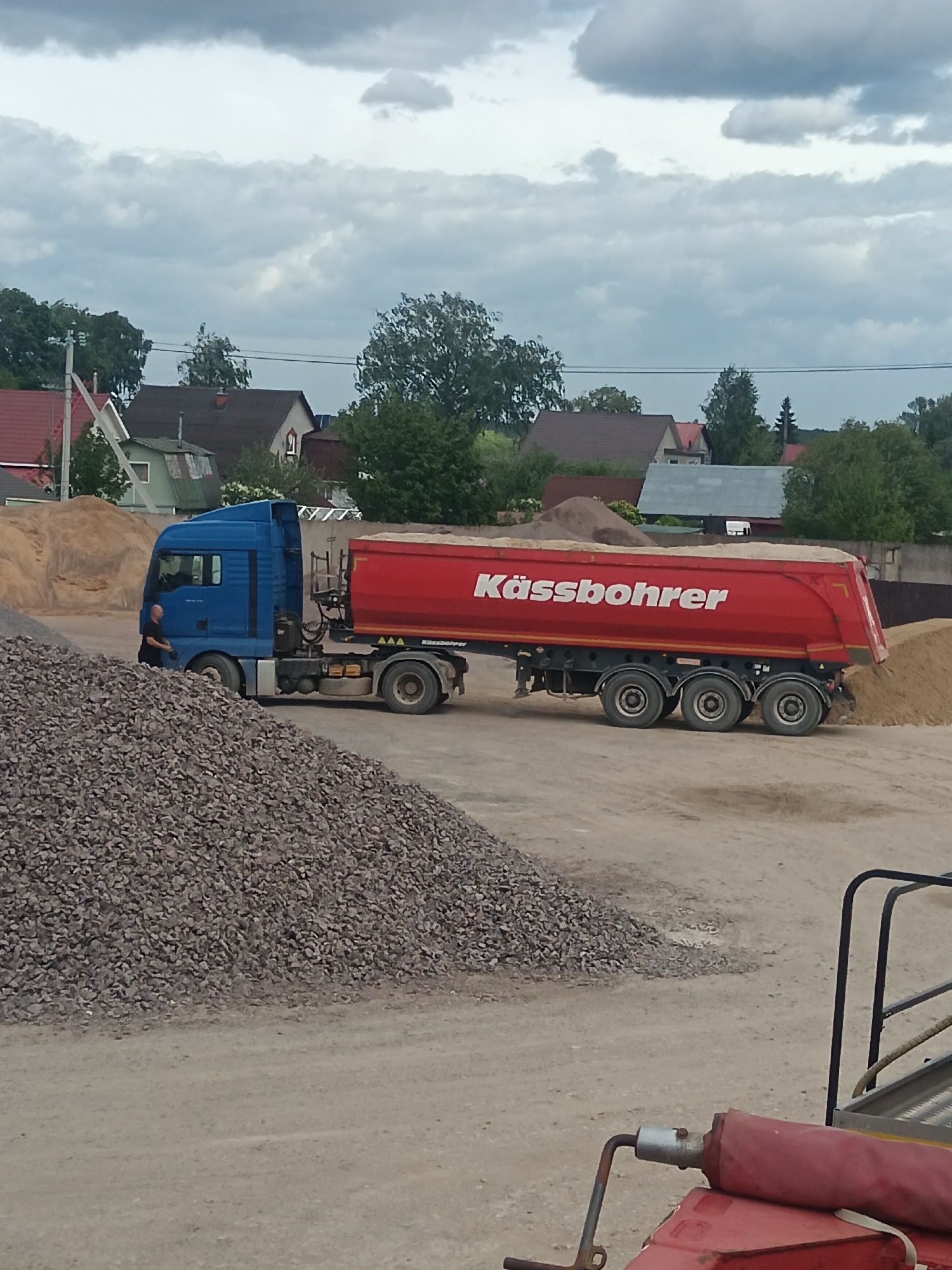 The image depicts a gravel pit or construction material storage site in Яльгелево, Russia. A blue semi-truck with a red trailer bearing the name "Kässbohrer" is the central subject, with the trailer filled with sand or gravel. A person is standing by the truck cab. There are large piles of gravel and sand visible in the foreground and background. The backdrop shows residential houses and trees under a cloudy sky.