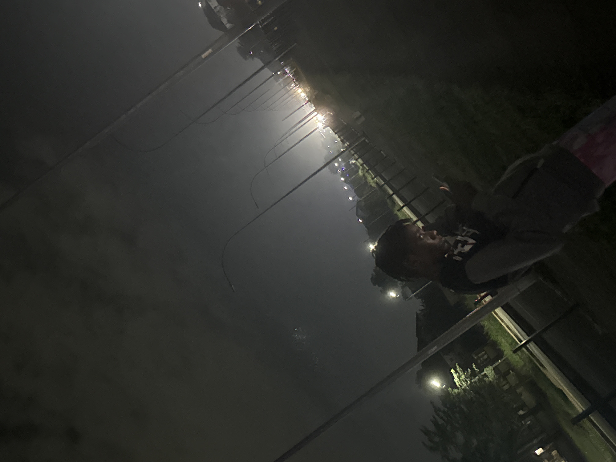 A young person is captured in a dimly lit outdoor scene, likely in the city of Uyo, Nigeria, at night. The individual is wearing a dark top over a lighter-colored garment and patterned trousers. They are holding a mobile phone and appear to be looking down at it.
The background is characterized by numerous streetlights lining what seems to be a road or pathway, creating a sense of depth and perspective. The lights are arranged in a receding pattern, suggesting an urban or semi-urban environment. Some of the lights in the distance are blurred, possibly due to the low-light conditions or atmospheric haze. Trees are visible on the left side of the image, their forms softened by the darkness. The ground is covered in grass, and a fence or railing is present in the mid-ground. The overall atmosphere is dark and slightly hazy, typical of an evening or nighttime setting in a location with ambient light pollution. The camera angle is slightly tilted, adding a dynamic feel to the composition.