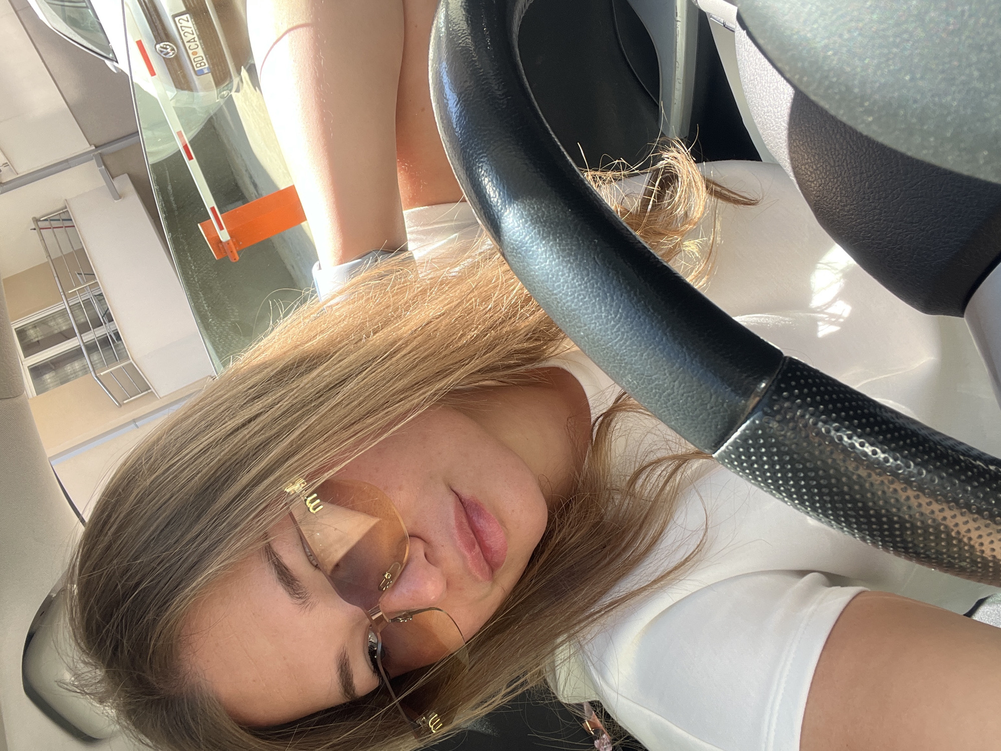 The image features a young woman resting inside a car, positioned so her head is visible in the foreground, facing towards the left side of the frame. Her long, light brown hair cascades around her face and over her shoulders. She wears light-tinted, gold-rimmed sunglasses with a distinctive "W" or "M" shaped detail on the temples, partially obscuring her eyes. Her lips are slightly parted, and her expression appears relaxed. She is wearing a white top, visible around her neck and shoulders.
A black, textured steering wheel dominates the right side of the image, partially covering the woman's upper body. Through the car's windshield, a reflection of the outside environment is clearly visible. This reflection includes a white car with a license plate reading "BD CA272," identifying the location as Budva, Montenegro (where "BD" is the regional code). In front of the reflected car, a white and red striped boom barrier with an orange lower arm is seen, suggesting an entry or exit point, possibly for a parking area or private residence. Further in the reflection, parts of a building with windows and a white, slatted structure are visible. Bright sunlight illuminates the scene, indicating it is daytime. The image captures a casual moment from inside a car, likely parked.