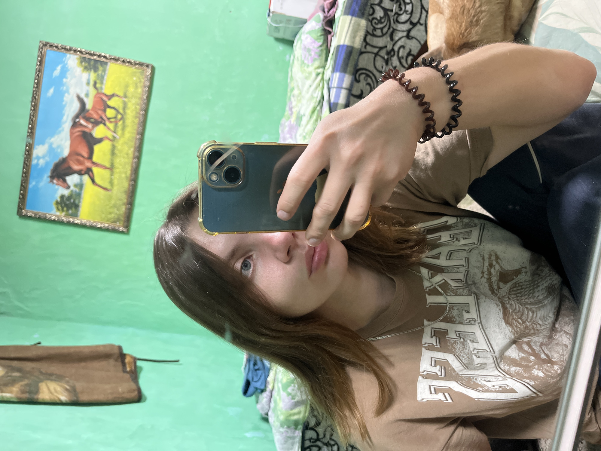A young person with long, light brown hair is taking a selfie in a room with bright green walls. They are wearing a tan t-shirt with a graphic print and a silver chain necklace. On their left wrist, they are wearing two spiral bracelets, one dark red and one black. The person is holding a dark-colored smartphone in their right hand, with the camera facing them. In the background, there is a framed picture of two horses on the wall, and a bed with patterned bedding. A piece of brown fabric hangs on the wall to the left of the person. The overall impression is that of a casual indoor snapshot.