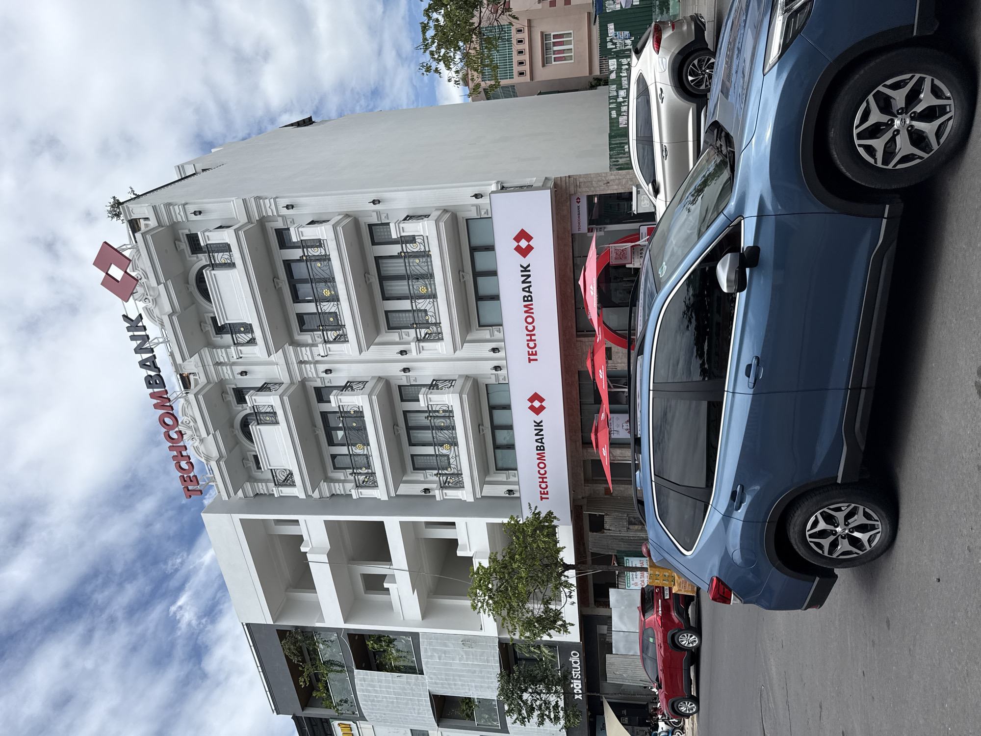 The image depicts a street view in Nha Trang, Vietnam, showcasing the Techcombank building. The building is a multi-story white structure with decorative balconies and the "TECHCOM Bank" logo prominently displayed in red and white. In the foreground, a blue Subaru Forester SUV is parked on the street, with a red SUV visible further down the road. The street appears to be a commercial area, with other buildings and signage also visible. The sky is partly cloudy, and trees are present along the street. No people are clearly discernible in the scene.