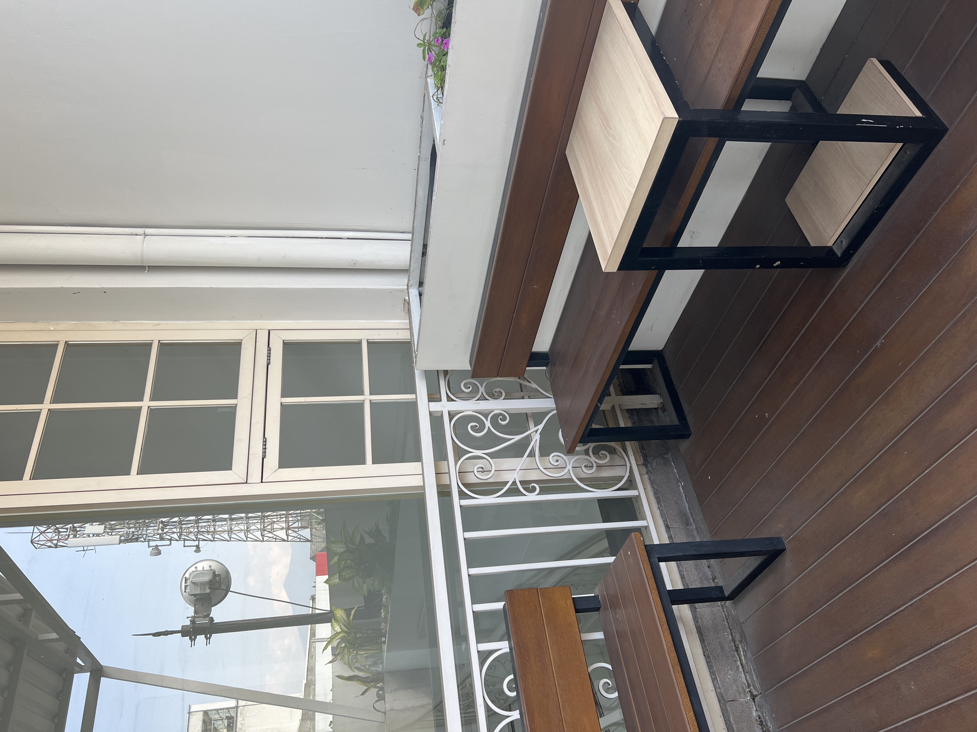 This is an eye-level outdoor shot of a seating area on a wooden deck, likely a balcony or patio, in Bandung City, Indonesia. The scene is dominated by two wooden benches with black metal frames, one of which is partially occupied by a small, light-colored wooden table also with a black metal frame. A white metal railing with decorative scrollwork separates the seating area from a lower level. Behind the railing, a large window with multiple panes reflects an outdoor scene, including a cell tower, a satellite dish, and distant mountains. The wall behind the benches is painted a light, neutral color and has two large, white pipes running vertically. In the background, a long, white planter box is visible, with some small pink flowers and green foliage. The wooden deck is laid with planks running diagonally from the bottom left to the top right of the frame. The overall impression is one of a quiet, functional outdoor space.