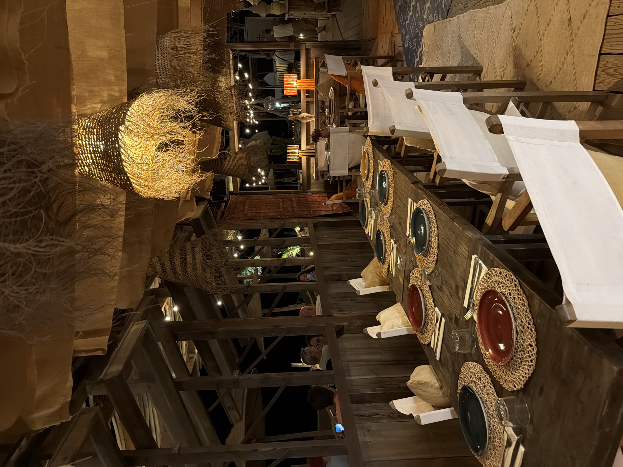 The image depicts the interior of a restaurant in Torreilles, France, characterized by a rustic and natural aesthetic. The scene is illuminated by warm, ambient lighting emanating from large, woven pendant lamps with frayed edges hanging from a beamed ceiling. The ceiling itself is draped with tan fabric, further contributing to the earthy ambiance.
In the foreground, a long, dark wooden table is set for dining. Each place setting includes a woven placemat, a red or dark green plate, silverware, and a glass. Several chairs with white canvas coverings are arranged around the table.
In the background, the restaurant's architecture is visible, featuring wooden beams and partitions. The restaurant appears to be open-air or have large openings, as evidenced by the view of the night sky with string lights and silhouettes of palm trees. Some patrons are visible in the background, seated at tables or standing. One person is holding a smartphone. The overall impression is that of a relaxed, beachside or tropical-themed dining establishment.