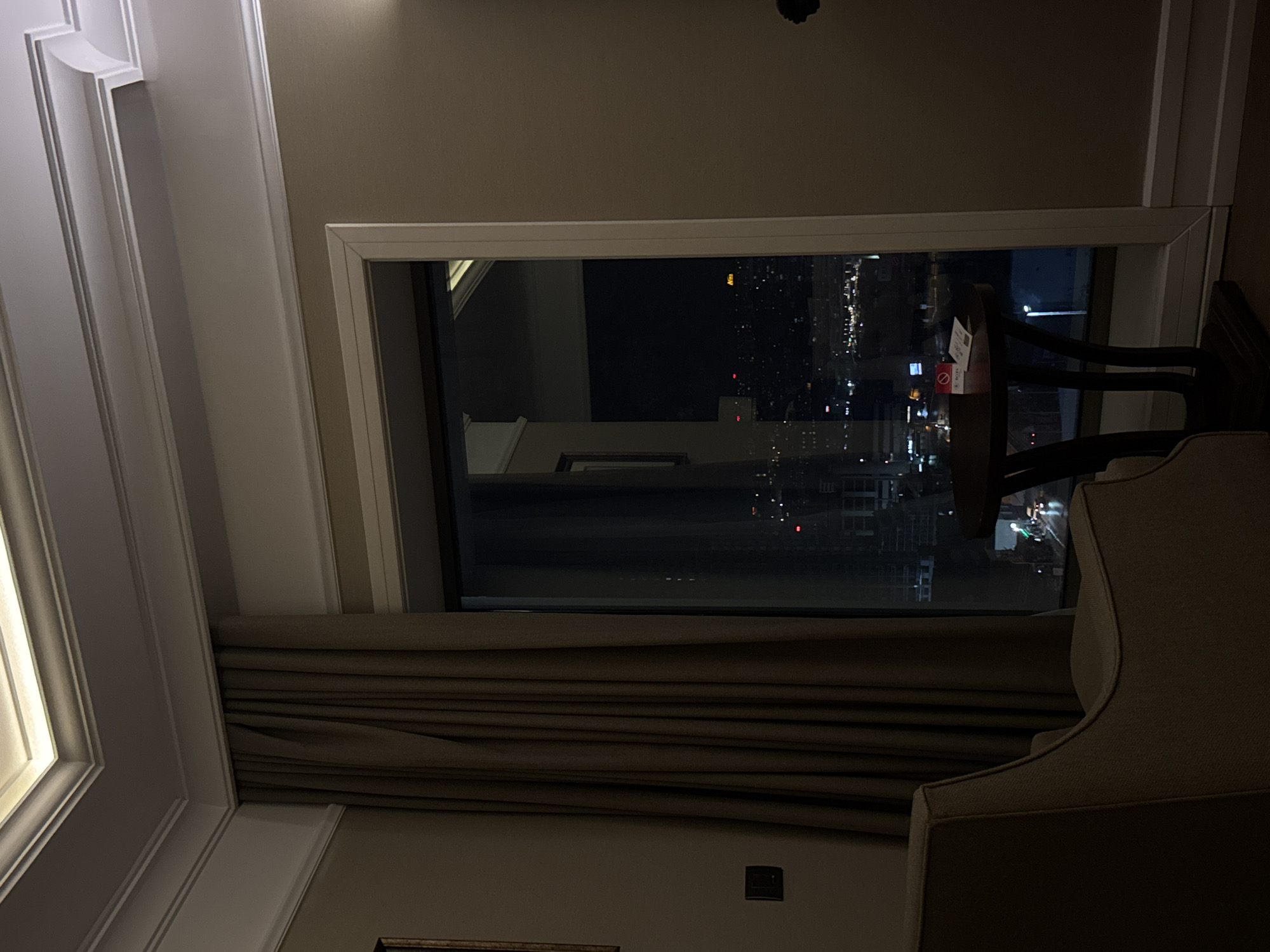 A view from inside a room overlooking a city at night. The room has curtains, a chair, and a table next to the window. The lights of the city are visible through the window. It is not possible to determine whether the image is of Bağcılar, Turkey based on the image content.