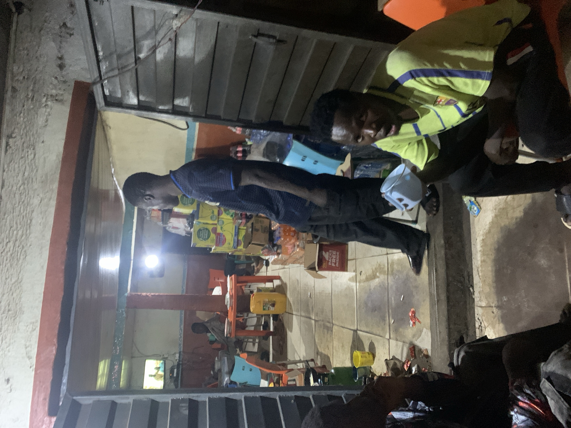 The image, captured in Gura Top, Nigeria, depicts an indoor commercial setting, likely a small shop or provision stall, illuminated by artificial light, suggesting it is nighttime.
Two men are prominently featured. In the central-left, a man wearing a dark blue collared shirt and dark trousers stands facing right, holding a white object, possibly a face mask. To the right, another man is seated or crouching. He wears a bright yellow and blue striped t-shirt, which appears to be an FC Barcelona jersey, and holds a white cup. He looks towards the standing man with a neutral to slightly smiling expression.
The background shows shelves stocked with numerous yellow-packaged goods or boxes. A cardboard box prominently displays the text "PACES FREE INSIDE." The shop's interior walls are painted in sections of beige, blue, and red, with a tiled floor that appears somewhat soiled in areas. Miscellaneous items such as plastic containers, a yellow jerry can, and scattered wrappers are visible on the floor. Orange plastic chairs and a small table are also present. The scene conveys a casual, everyday interaction within a functional local retail environment.