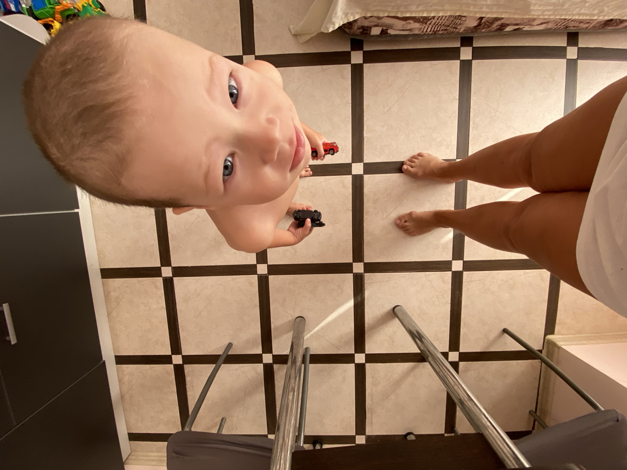 This image depicts a child and an adult from a high-angle perspective, seemingly indoors. The child, the primary subject, is positioned in the center of the frame and is looking upwards. They have blue eyes and fair skin, with light brown hair. The child is holding two small toy cars, one red and one black. To the right of the child, an adult's legs and feet are visible, suggesting they are standing or sitting on the floor.
The scene is a room with a tiled floor, characterized by a grid of light-colored square tiles with dark grout lines. In the background, a bed with patterned bedding is partially visible. There are also metal chair legs and parts of chairs in the foreground, suggesting a dining or living area. To the left, a dark piece of furniture, possibly a cabinet or door, is visible.
The lighting suggests it might be daytime, but there are no specific indicators of the time of day or weather conditions. The overall mood appears to be casual and domestic, possibly a moment of play or interaction between the child and adult. The location context provided is Sochi, Russia, though no specific landmarks or cues within the image directly confirm this. No visible text is present in the image.