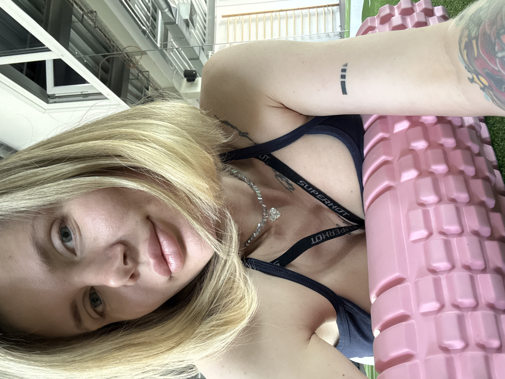 A young woman with blonde hair and light blue-grey eyes takes a close-up selfie while lying down, possibly on an exercise mat, in an indoor gym in Mayma, Russia. She looks directly at the camera with a neutral, slightly relaxed expression. Her blonde hair frames her face, and her lips appear full. She wears a dark sports bra with white text that reads "SUPERHOT" visible on the straps. A sparkling necklace with a large, clear pendant adorns her neck. Distinct tattoos are visible on her arms: a small, abstract black mark resembling an exclamation point or lowercase 'i' on her left inner bicep, and a larger, colorful tattoo on her right arm, partially obscured. Beside her, a large, pink foam roller with a textured, knobbed surface is prominently placed, resting on what appears to be artificial green turf. The background reveals an industrial-style gym ceiling with exposed metal beams, ventilation ducts, and bright overhead lighting. A large window or skylight structure is also part of the ceiling design, contributing to the well-lit environment.