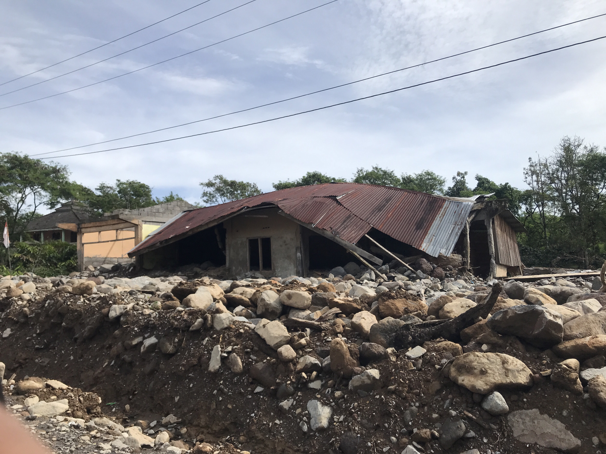 The image shows a severely damaged building in what appears to be a disaster-stricken area in Bintangpermata, Indonesia. The house has a partially collapsed corrugated metal roof and concrete walls, indicating structural compromise. A field of rocks and debris surrounds the base of the building, suggesting a mudslide or flash flood event. In the background, other buildings are visible, though their condition is not immediately clear. The sky is overcast. Overhead power lines are visible in the sky. No people are visible in the frame. The scene suggests the aftermath of a natural disaster event.