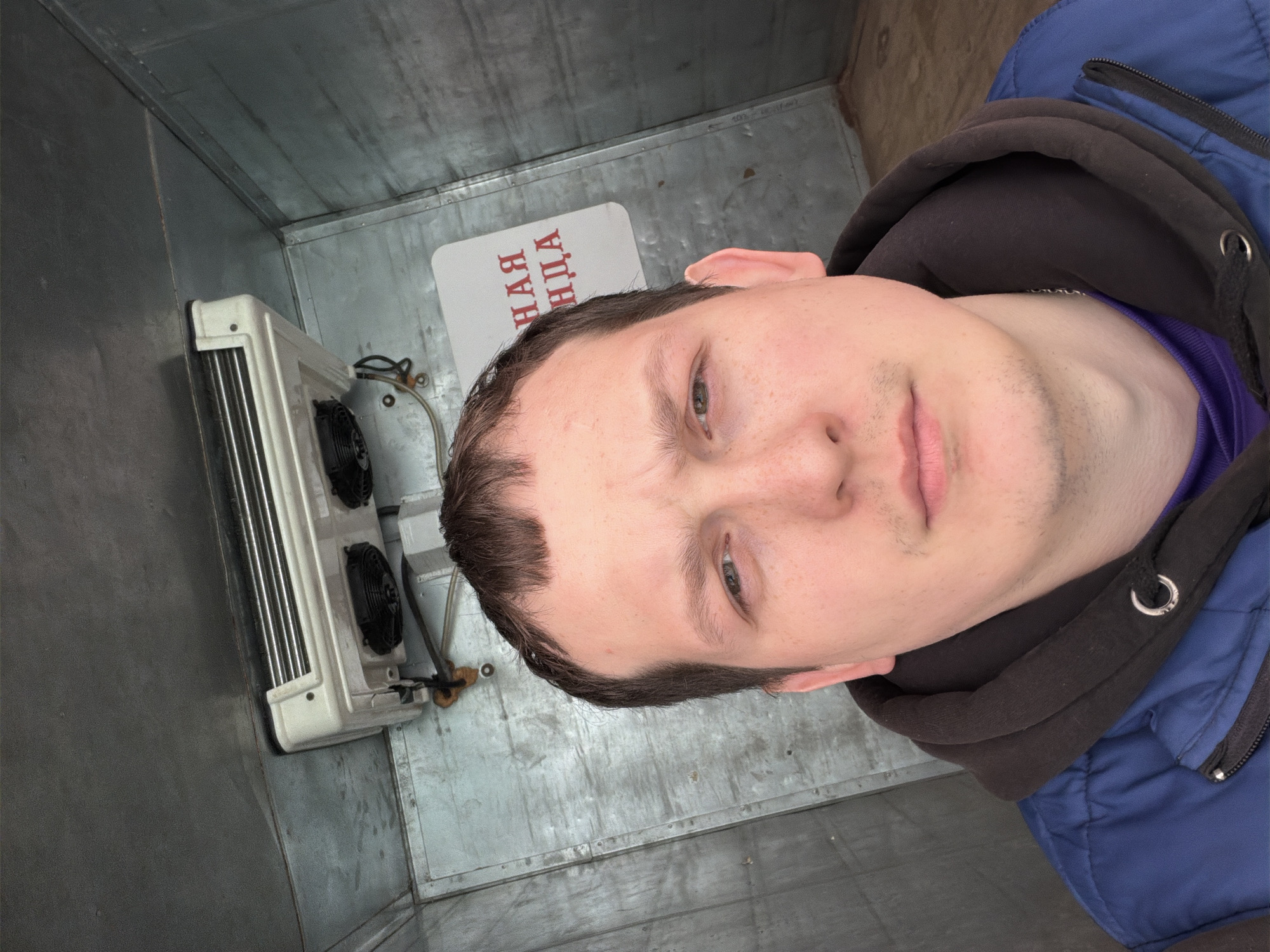 A medium, eye-level, portrait-style shot shows a person inside what appears to be a refrigerated storage unit or truck. The person is light-skinned, with short brown hair, and wearing a blue jacket with a dark hoodie and a purple shirt visible at the neck. The face is angled slightly to the right of the frame, and the person has a neutral expression.
To the left, a white refrigeration unit with two black fans is mounted on a gray, metallic wall. A sign in Cyrillic script is mounted on the wall to the right of the refrigeration unit. The background consists of gray, metallic walls and ceiling, suggesting an enclosed space.
Given the presence of Cyrillic script on the sign and the information provided, this image is likely taken inside a refrigerated unit in the city of Chaikovsky, Russia.