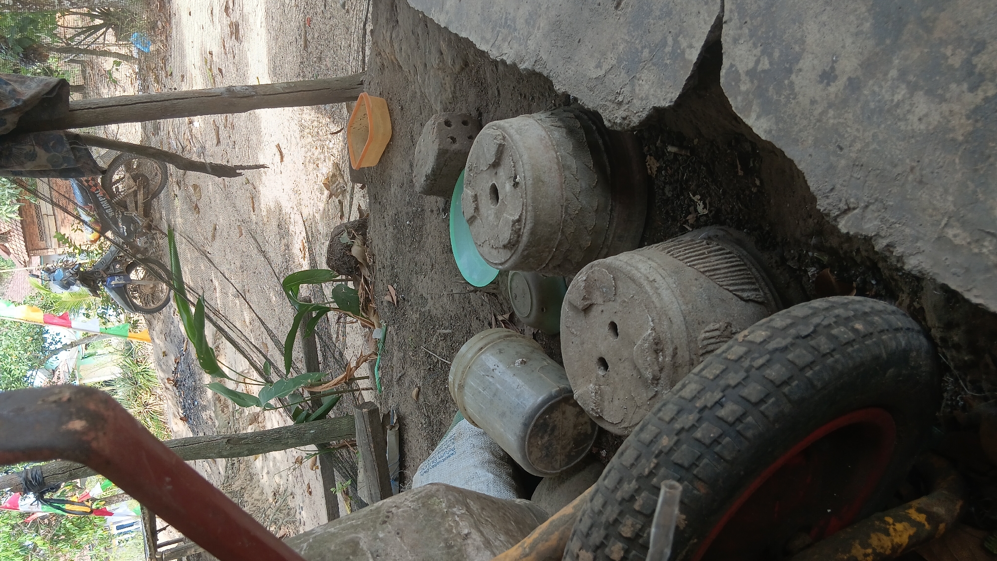 The image depicts an outdoor scene in Prabumulih, Indonesia, characterized by a somewhat unkempt, dusty environment with various objects scattered around. In the foreground, a large, dark-colored tire with a red rim is prominently visible, partially obscuring a pile of concrete pots, one of which has two holes on its surface. To the left of the pots, a large, greyish, cylindrical object, possibly a bag or a concrete structure, is present, along with a clear plastic container lying on its side. Behind these objects, a green plastic saucer rests on the ground, and further back, another concrete pot with intricate carvings is seen. An orange plastic container is also visible in the mid-ground.
The background reveals a dirt path or ground, with a motorcycle parked in the distance. Several flags, including red, white, and green ones, are affixed to poles, suggesting a possible celebration or event. A structure with a tiled roof and brick walls is partially visible, indicating a residential or storage area. Green foliage and plants are scattered throughout the scene, and a thin layer of netting covers part of the area, possibly for protection or containment. The overall impression is one of an outdoor storage or working area, possibly on the outskirts of a residential zone in Prabumulih.