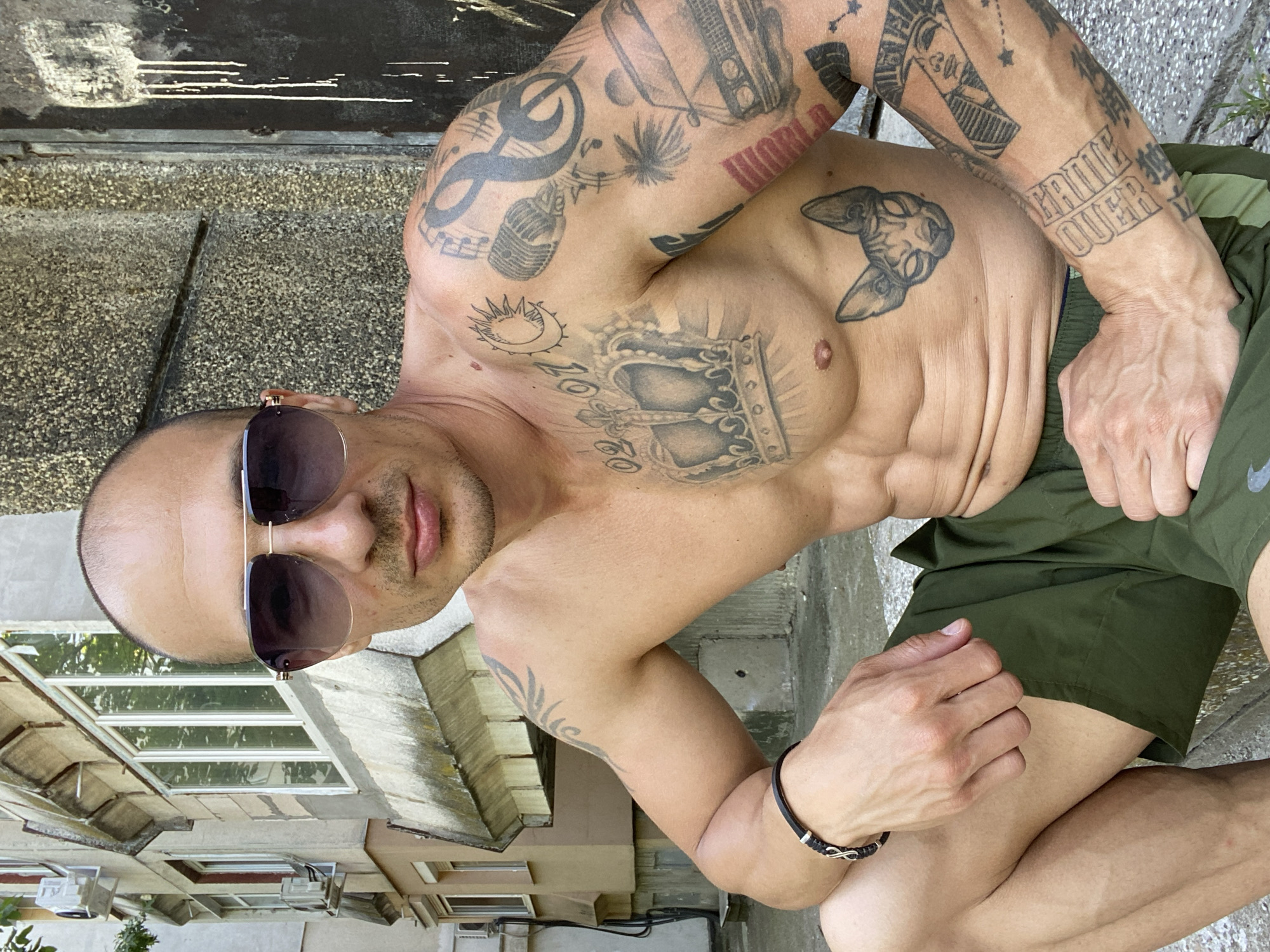 A bare-chested man with a bald head, sunglasses, and extensive tattoos on his torso and arms is crouching outdoors. He is wearing olive-green shorts and a black bracelet on his left wrist. The background features a concrete wall with peeling paint and a weathered building with balconies and windows. The scene appears to be in an urban setting, possibly in Pleven, Bulgaria, given the architectural style. The man's pose and expression are relaxed, and he is looking towards the camera.