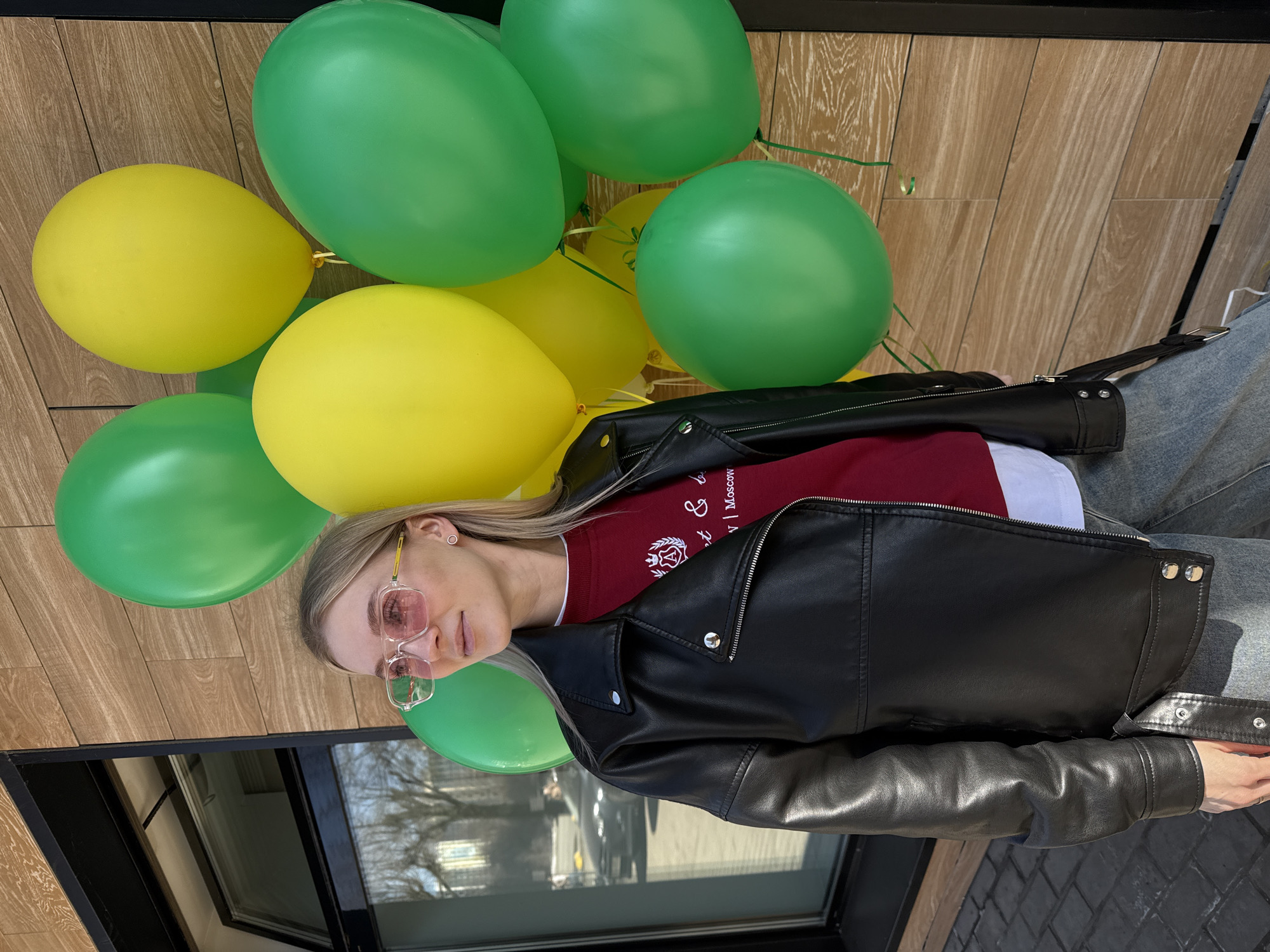 A young woman with blonde hair and pink sunglasses is holding a bunch of green and yellow balloons. She is wearing a red and white sweatshirt under a black leather jacket and blue jeans. The background features a wooden wall and a window reflecting a city street. The scene is likely taking place outdoors, possibly in Izhevsk, Russia, judging by the lettering on the sweatshirt.
