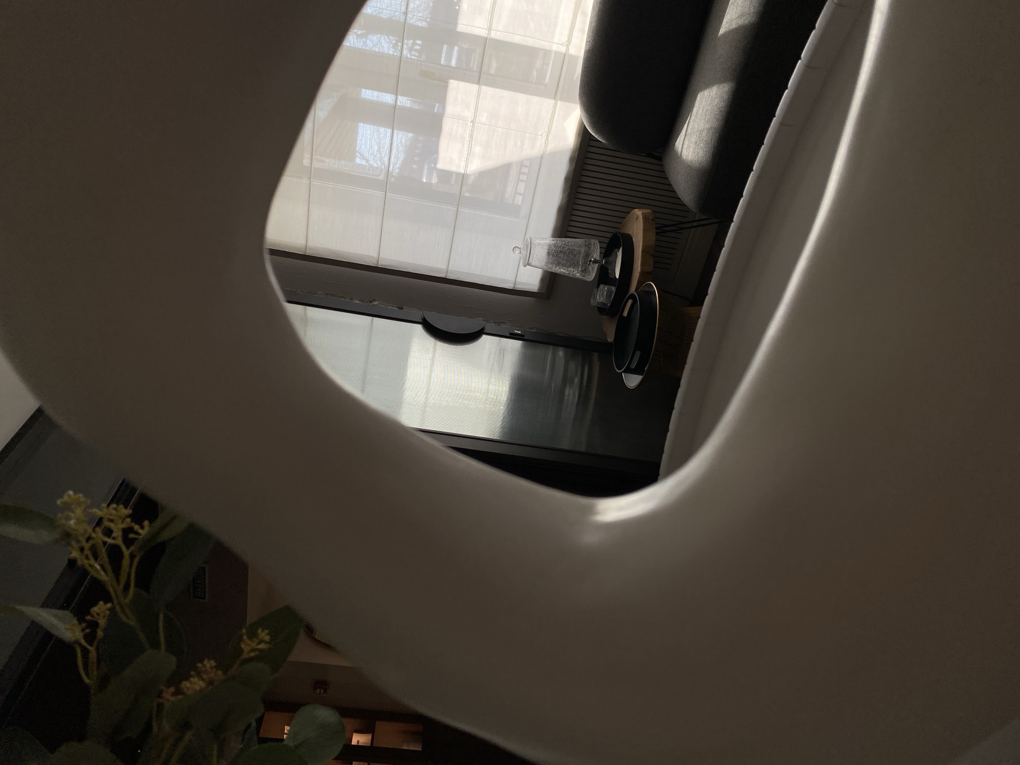 The image, taken with a portrait orientation and rotated 90 degrees clockwise, captures an interior scene through the organic curve of a large, off-white object, possibly a modern furniture piece or architectural element.
In the upper portion of the frame (when viewed upright), a large window dominates, fitted with translucent white horizontal blinds or shades. Bright, diffused sunlight streams through the window, casting reflections and soft shadows. Beyond the window, hints of an outdoor scene with trees or buildings are visible but indistinct due to the blinds and glare.
Below the window is a dark, reflective surface, likely a television screen or a polished tabletop. To the right of this surface, on what appears to be a small side table, are several items: a clear glass carafe or decanter, a small circular wooden object (possibly a tray or coaster), and two dark, circular objects that could be plates or decorative accents. To the right of these items, a portion of dark gray upholstered furniture, possibly an armrest of a sofa or chair, is visible.
In the lower-left corner, a green plant with numerous small, light yellow or off-white flowers or buds adds a touch of nature to the indoor setting. The lighting suggests it is daytime, likely a bright, sunny day. The scene is devoid of people and activity, presenting a calm, static snapshot of an interior space. Very faint, unreadable white text is barely visible on a dark surface in the distant background. The location context for this scene is Votkinsk, Russia.