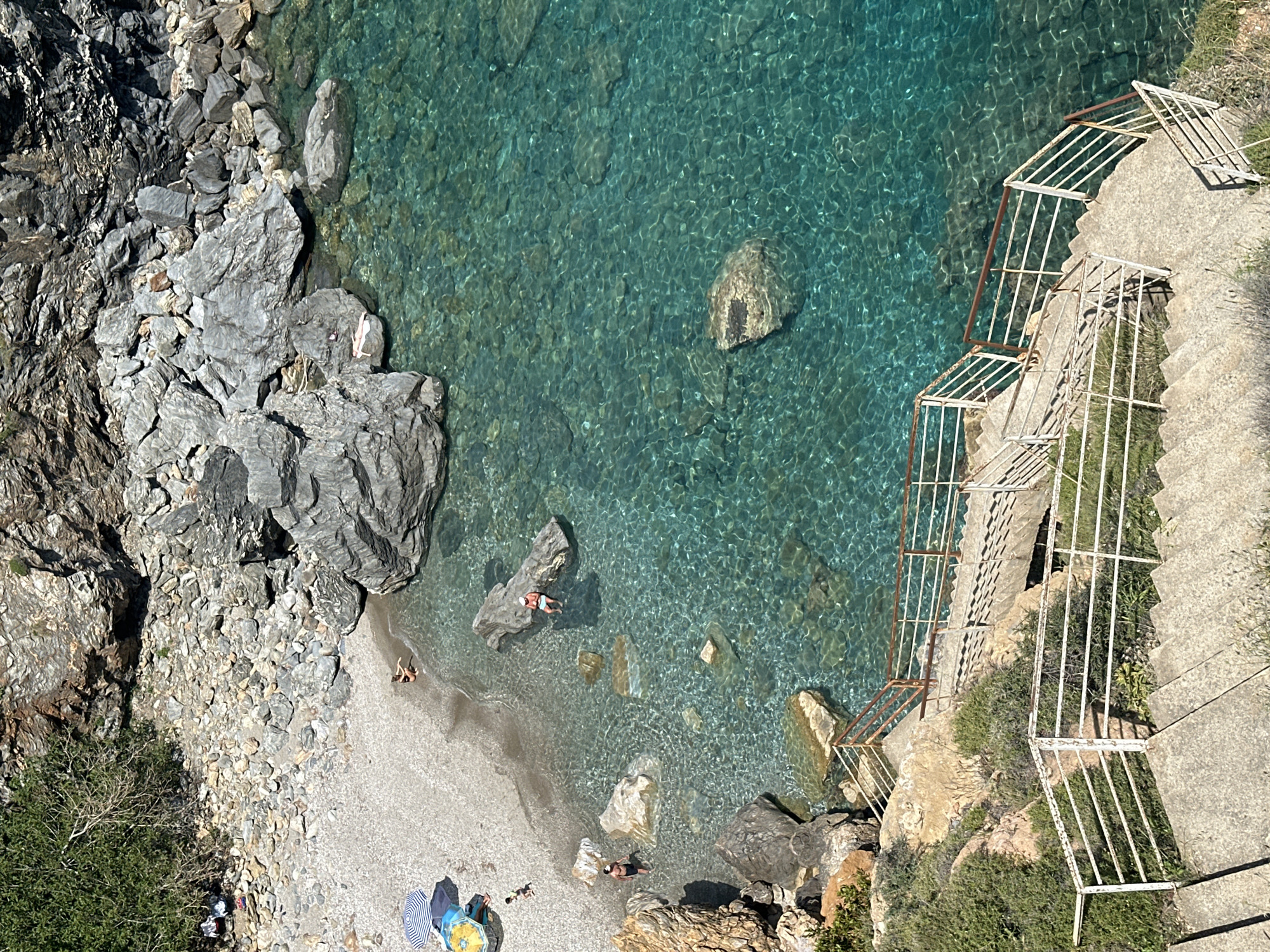 An aerial view looking down at a small beach cove in Gazipaşa, Turkey. Clear turquoise water fills the cove, revealing rocks beneath the surface. A rocky shoreline borders one side, while concrete steps with a metal railing descend from a cliff to the beach on the opposite side. Several people are visible: one swimming, a few lying on the beach, and others near a beach umbrella. Rocks and pebbles cover the beach area. Vegetation grows on the cliffside.