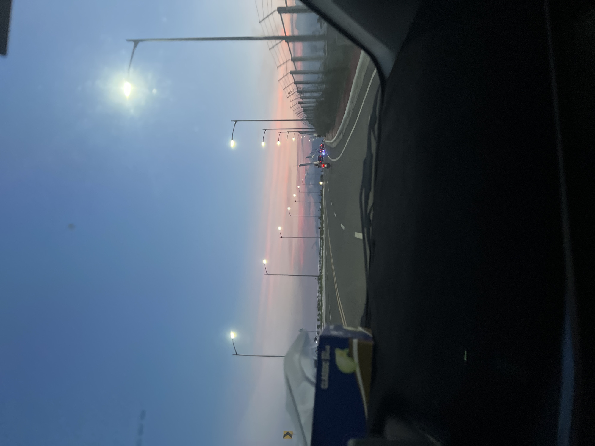 The image presents a candid view from inside a vehicle, looking straight ahead on a multi-lane asphalt road at dusk or sunset in Davao City, Philippines. The sky transitions from a deep blue overhead to vibrant hues of pink, orange, and purple near the horizon, indicative of twilight. Numerous modern streetlights with bright white illumination line both sides of the road, casting light on the scene.
In the immediate foreground, a blue box of "CLASSIC 50s" tissues rests on the dashboard, partially obstructing the view of the left side of the road. The road itself features clear white lane markings and extends into the distance, appearing relatively clear of heavy traffic.
On the left side of the road, a yellow chevron sign indicating a curve or turn is visible. Further along, a low barrier or railing separates the road from the unseen landscape beyond. On the right side of the road, a tall chain-link fence topped with barbed wire runs parallel to the roadway, strongly suggesting a security perimeter. Beyond this fence, indistinct silhouettes of trees or distant structures are visible.
In the mid-ground, several vehicles, including what appear to be motorcycles and cars, are traveling ahead on the road, distinguishable by their illuminated headlights and taillights. The overall environment suggests an urban or peri-urban highway.
Forensically, the presence of a multi-lane highway with modern infrastructure and a distinctive security fence points to a location adjacent to the Francisco Bangoy International Airport (DVO) in Davao City, Philippines, likely a section of the Davao City Diversion Road or Pan-Philippine Highway that runs along the airport's perimeter.