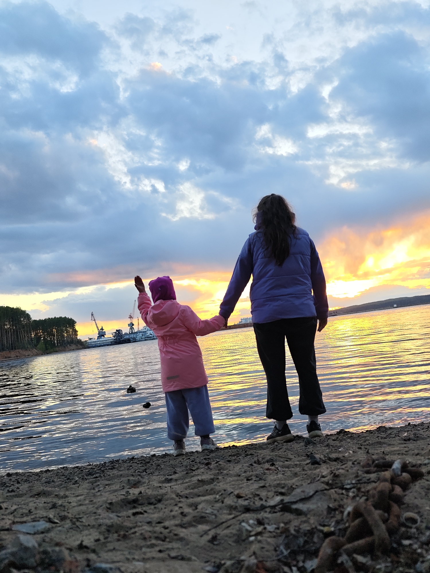 The image shows a woman and a child standing on a sandy shore near a body of water, likely the Kama River. The location is likely Chaikovsky, Russia, based on the presence of industrial structures like cranes and ships visible in the background. The sky is filled with clouds during a sunset, creating a golden reflection on the water. The child, wearing a pink coat and purple hat, has one arm raised. The woman, dressed in a purple jacket and dark pants, holds the child's hand. A segment of a rusted chain is visible in the foreground on the beach.