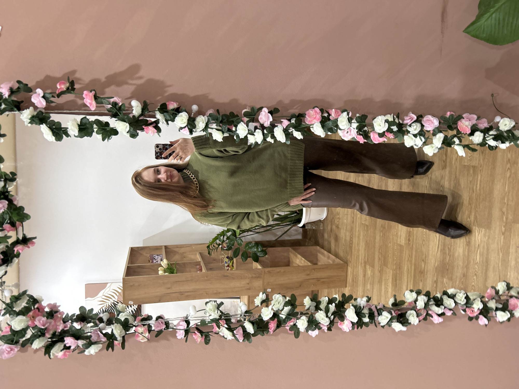 A woman is standing in front of a mirror, taking a selfie with her phone. She is wearing a green sweater and brown pants. A wooden shelf is visible in the mirror's reflection, along with plants. The mirror is decorated with a garland of pink and white flowers. The location is likely an interior space in an unknown location, possibly Chaikovsky, Russia.
