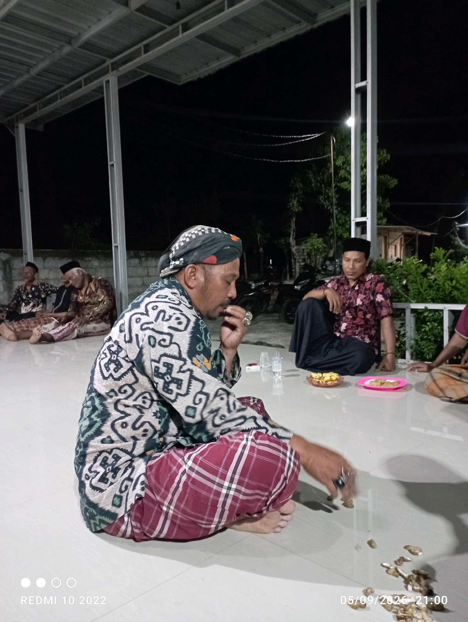 The image depicts a nighttime gathering of at least five men in what appears to be an outdoor covered area, likely in Dukuhseti, Indonesia, given the date stamp "05/09/2026 21:00". The setting is illuminated by artificial light, casting shadows. The ground is a light-colored tiled surface.
In the foreground, a man wearing a distinctive patterned robe and a headscarf is seated on the floor, with his right hand near his mouth, possibly eating or smoking. His legs are crossed, and he is looking down. He wears a ring on his right index finger.
To his left and further back, two older men are also seated on the floor. The man on the left wears a dark patterned shirt and a peci (cap). The man in the middle wears a patterned robe and a peci.
To the right of the foreground man, another man is seated with his legs crossed. He wears a patterned short-sleeved shirt and dark trousers. On the floor in front of him are a bowl of bananas and a pink plate with some food items.
Further to the right, part of another person is visible, also seated on the floor.
The background shows a dark night sky, trees, and some structures, including motorcycles parked behind the seated men. A metal roof structure supported by white pillars is visible overhead, suggesting an open-sided building or a veranda. The overall atmosphere appears to be informal and communal.
