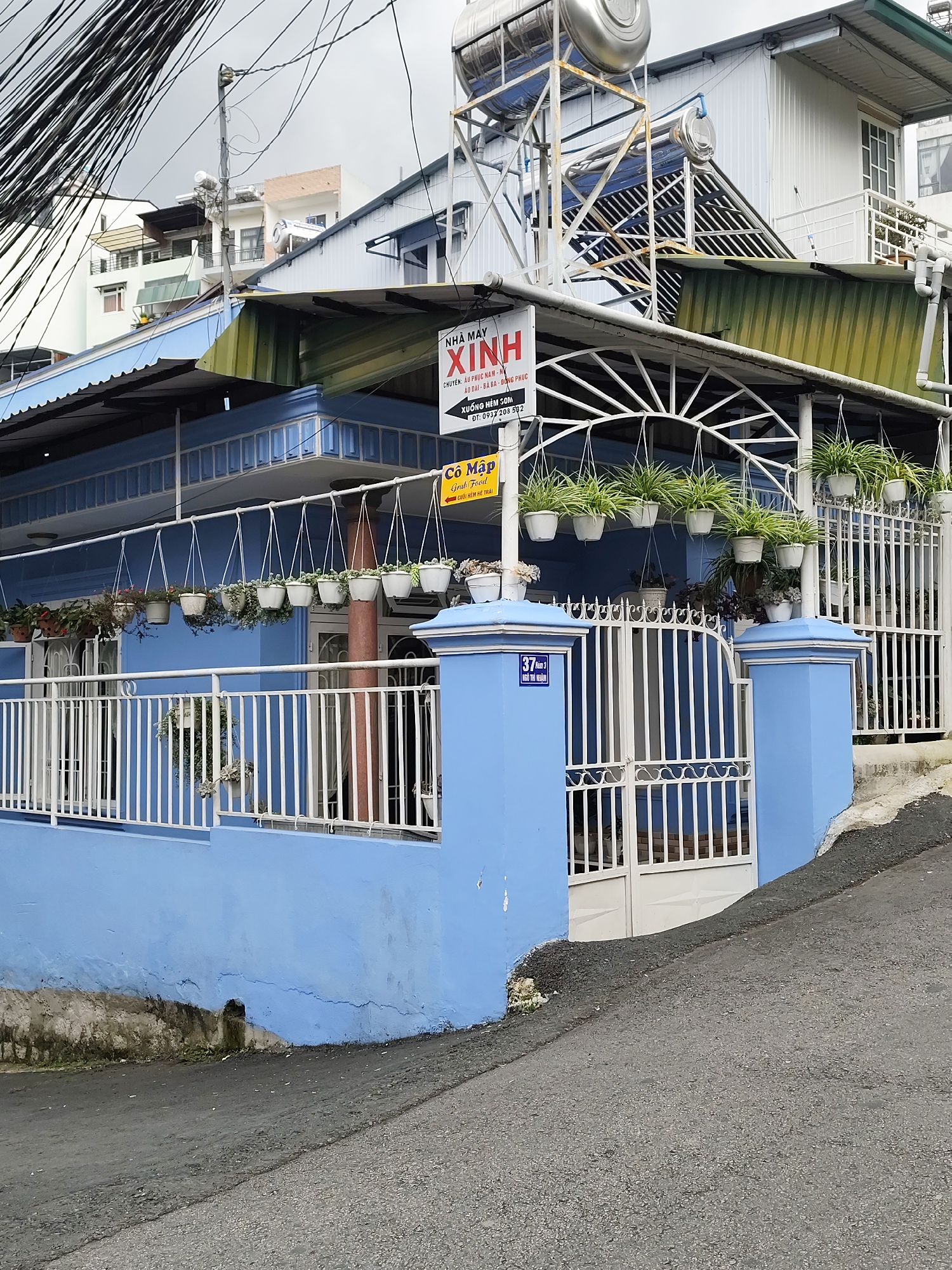 The image depicts a residential building in Da Lat, Vietnam. The building is painted light blue and has a white metal fence in front. Several hanging potted plants are visible on a white metal rack above the fence. A sign with Vietnamese text is attached to the building. The street in front of the building slopes upwards, and there are power lines overhead. The sky is overcast. The building appears to be a single-family home or a small commercial establishment.