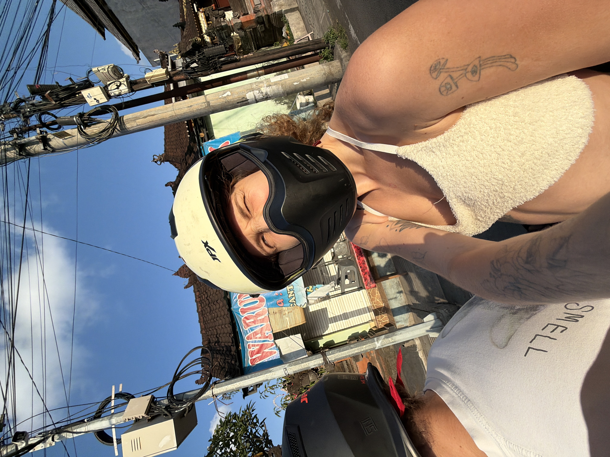Two individuals are riding a motorcycle on a street in the city of Mengwi, Indonesia. The person in the foreground is wearing a white and black helmet, a white fuzzy top, and has a mushroom tattoo on their right arm. The person in the background is wearing a black helmet and a white t-shirt with the word "SMELL" printed on it. The background shows a building with traditional Balinese architecture, a street sign that reads "WARUNG PANTURA," and numerous utility poles with tangled wires. The sun is shining, casting shadows on the ground.
