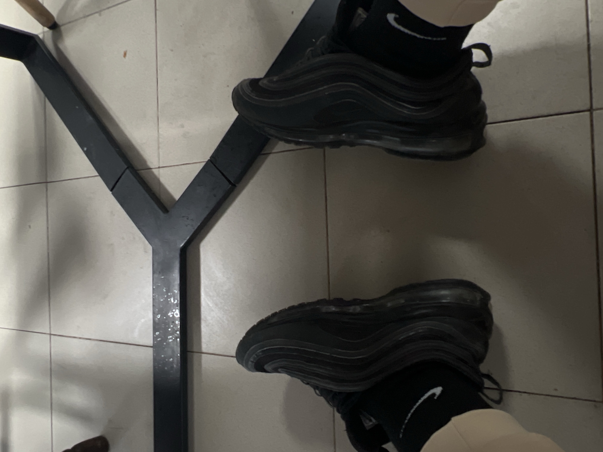 The image shows a first-person perspective of a person wearing black Nike Air Max 97 sneakers and black socks with the Nike swoosh logo. The person's beige pants are also visible. The shoes are placed on a tiled floor next to a black Y-shaped metal table leg. The location is unknown and there is no indication that the photo was taken in Goce Delcev, Bulgaria.