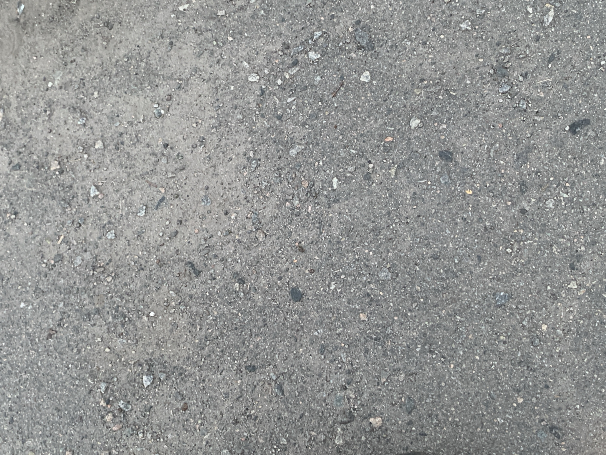 The image shows a close-up view of a gray asphalt surface with small stones and gravel embedded in it. The texture is rough and uneven, typical of a road or pavement. There are no people or specific objects visible, just the ground itself. The lighting suggests it is daytime, with a diffused light casting no strong shadows. The scene is devoid of any context that would indicate a specific location within a city or otherwise.