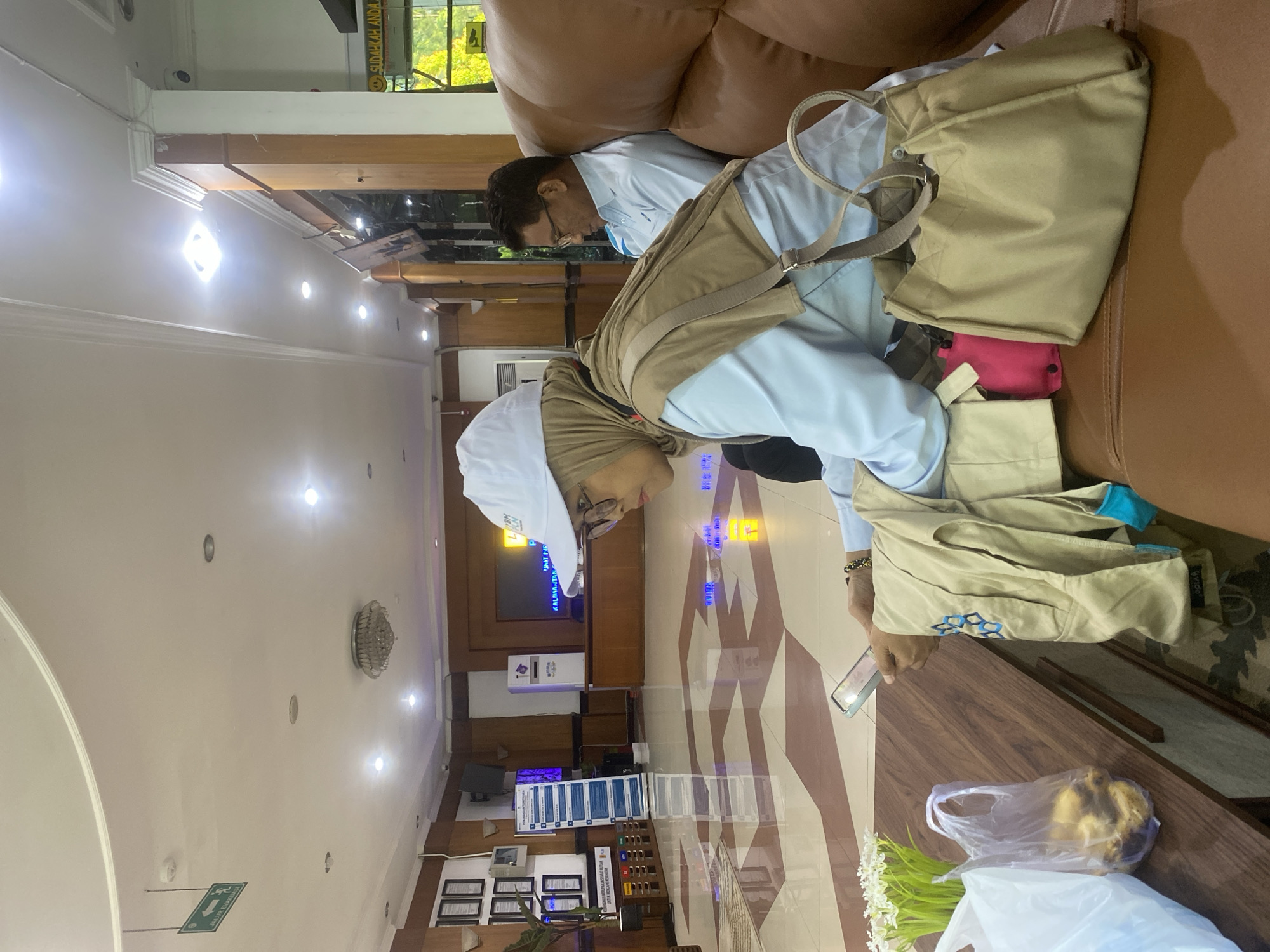 This image was taken in the waiting area of the PLN office in Banjarbaru, Indonesia. The scene depicts two individuals seated on a brown leather sofa. In the foreground, a woman wearing a light blue long-sleeved shirt, a beige hijab, and a white baseball cap is engrossed in her smartphone. She has a beige vest or jacket draped over her lap, and a beige handbag is placed beside her on the sofa. To her left, on a wooden table, are a plastic bag containing what appear to be pastries, a bouquet of white flowers, and another plastic bag. Behind the woman, a man wearing a light blue collared shirt and glasses is seated, looking down. The background reveals the interior of an office waiting area, characterized by a patterned tiled floor, wooden paneling, and overhead lighting. A large screen displaying text is visible on the wall. An emergency exit sign is also mounted on the ceiling.