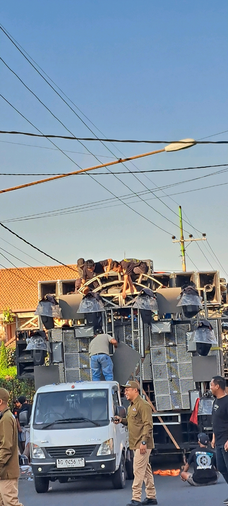A group of people are working on a large sound system mounted on a truck in the city of Prigen, Indonesia. The sound system consists of numerous speakers and is housed in a metal frame. Several individuals are positioned on top of the sound system, appearing to be adjusting or securing equipment. Below them, another person in blue jeans and a grey shirt is also working on the sound system. In the foreground, a white truck with the license plate "AG 9456 VG" is parked, and a man in a brown uniform and cap is smiling and holding a phone. Other individuals are visible in the background, and power lines crisscross the sky. The scene suggests preparation for an event or performance.