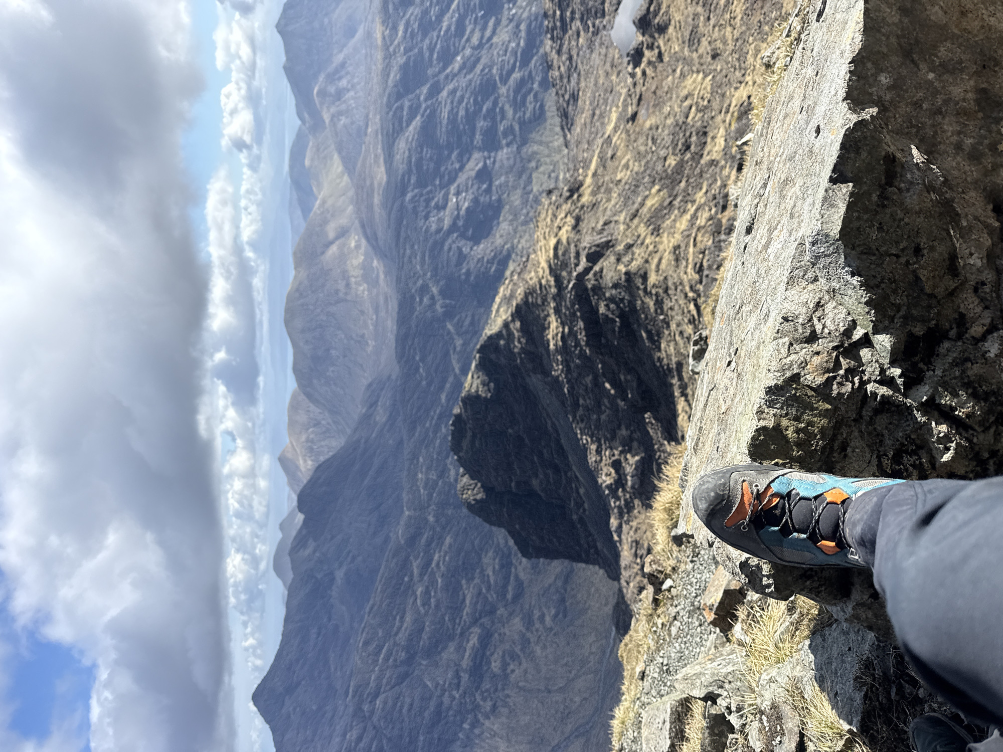 A high-angle, wide shot displays a climber's foot in a climbing shoe poised on a rocky ledge overlooking a mountainous landscape. The right side of the frame shows the climber's foot, clad in a grey climbing shoe with teal and orange accents. The leg, covered in dark grey pants, extends partially into the lower right corner. The foot is situated on a craggy, light grey rock ledge. Beyond the ledge, the vista opens up to reveal a deep valley and a series of rugged, dark-colored mountains. In the background, a cloudy sky stretches across the horizon, transitioning from darker clouds on the left to lighter patches of blue and white. The landscape suggests a remote, mountainous region. The city is unknown.
