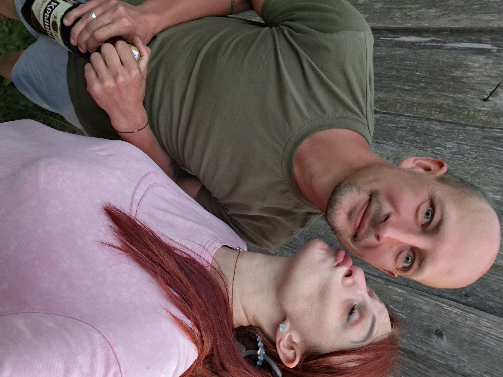A man and a woman are posing for a photo against a weathered wooden wall, presumably in Забалоцкі сельскі Савет, Belarus. The man, bald with a goatee, wears a green t-shirt and holds a beer bottle. The woman, with long red hair, leans in to kiss him, wearing a pink t-shirt and heart-shaped earrings. They appear to be outdoors, with hints of grass visible in the foreground.