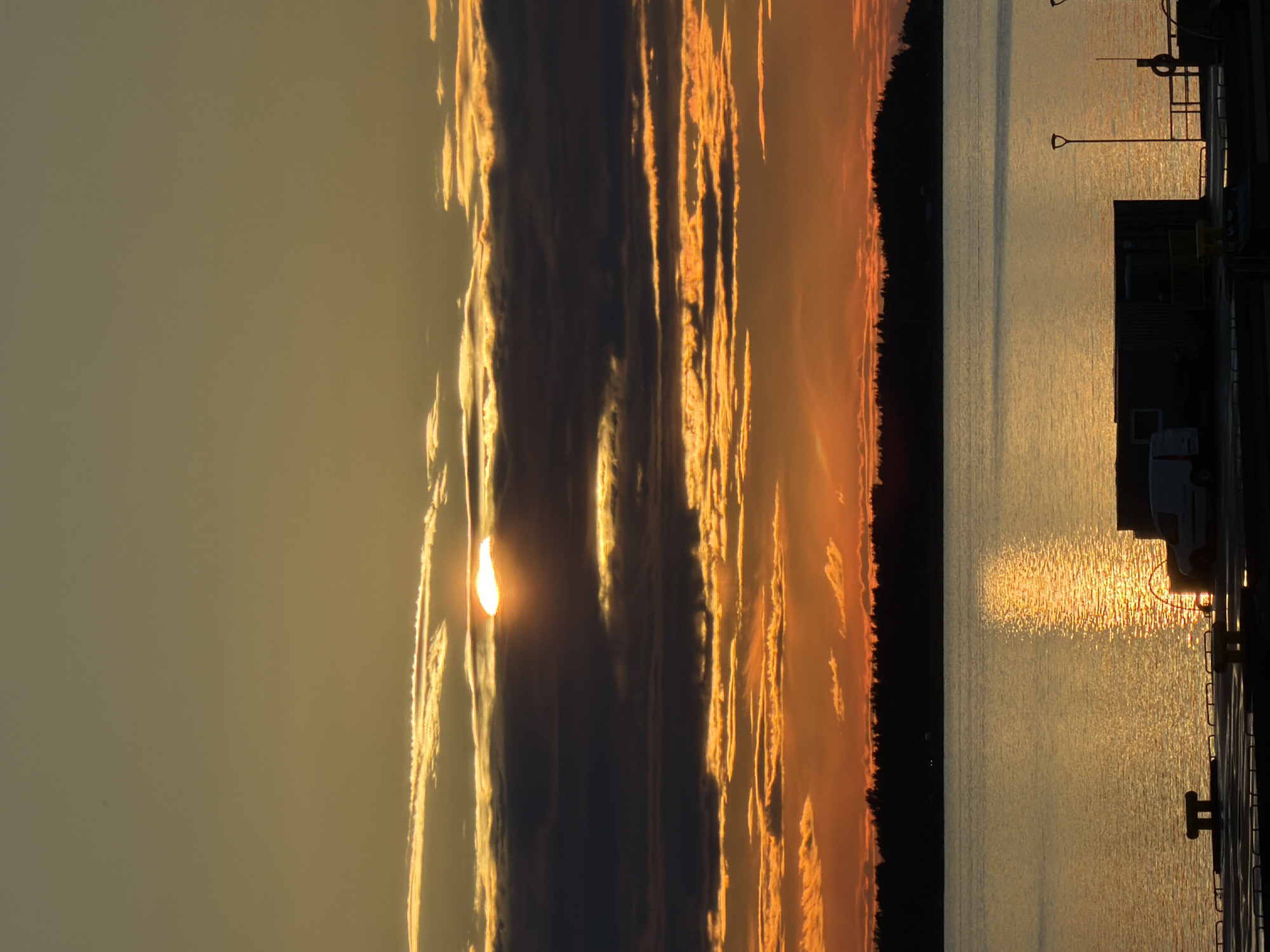 The image depicts a serene sunset over the Baltic Sea, likely near Tallinn, Estonia, as indicated by the silhouetted treeline resembling the coast and the presence of port-like structures. The sky is ablaze with warm hues of orange and yellow, with the sun peeking through a layer of dark clouds, casting a golden reflection on the calm water. In the foreground, a pier is visible with a white van parked near a container-like structure. The overall atmosphere is peaceful and tranquil, characteristic of a coastal sunset.