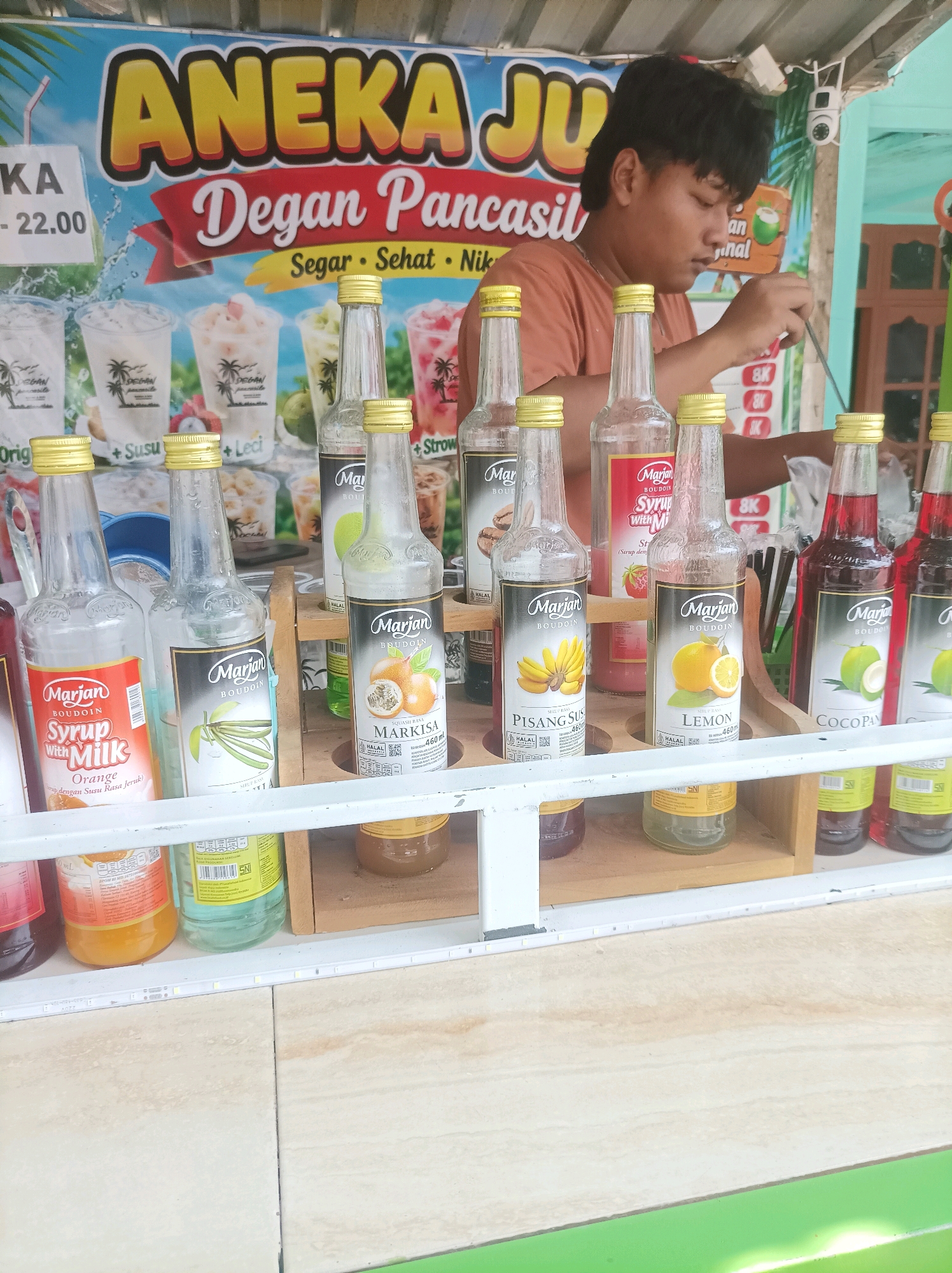 The image shows a street food stall in Sumuragung, Indonesia, selling various flavors of Marjan Boudoin syrup. A young man, likely the vendor, is seen behind the counter, preparing a drink. The stall displays an array of bottled syrups, including flavors like "Syrup with Milk Orange," "Markisa," "Pisang Susu," and "Lemon." The stall also advertises "Aneka Jus Degan Pancasil" (Assorted Coconut Pancasil Juice) and mentions prices such as "22.00." In the background, there's a banner with colorful illustrations of drinks and fruits. The overall environment appears to be an outdoor setting, typical of street food vendors in Indonesia.