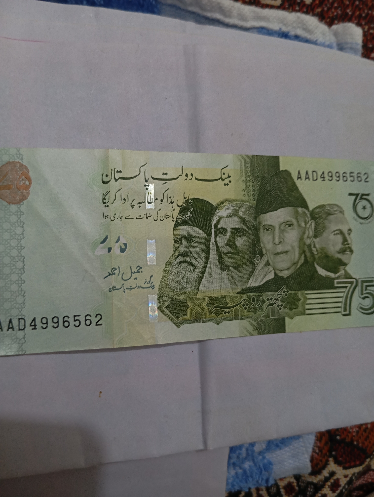 The image shows a close-up of a Pakistani 75 Rupee banknote. The banknote is predominantly green and features portraits of prominent Pakistani figures. From left to right, the figures visible are possibly a religious leader, a woman, Muhammad Ali Jinnah, and possibly another leader. The denomination "75" is clearly visible in the lower right corner, both numerically and in Urdu script. The serial number "AAD4996562" is also present in the upper right and lower left corners. The banknote appears to be in good condition, with some minor creases visible. The background is out of focus and appears to be a textured surface. No specific location within a city is discernible from the image.