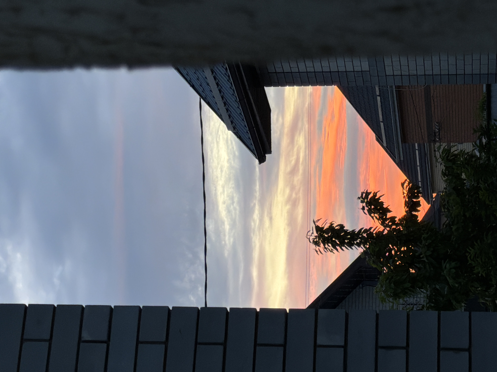The image shows a sunset sky viewed from what appears to be between buildings or through a gap in a wall. The sky transitions from a cloudy grey on the left to vibrant orange and yellow hues on the right. Parts of buildings and some greenery frame the colorful sky. The buildings appear to be residential. The location is given as Rossiyskiy, Russia, though there are no specific clues in the picture itself to confirm this location.